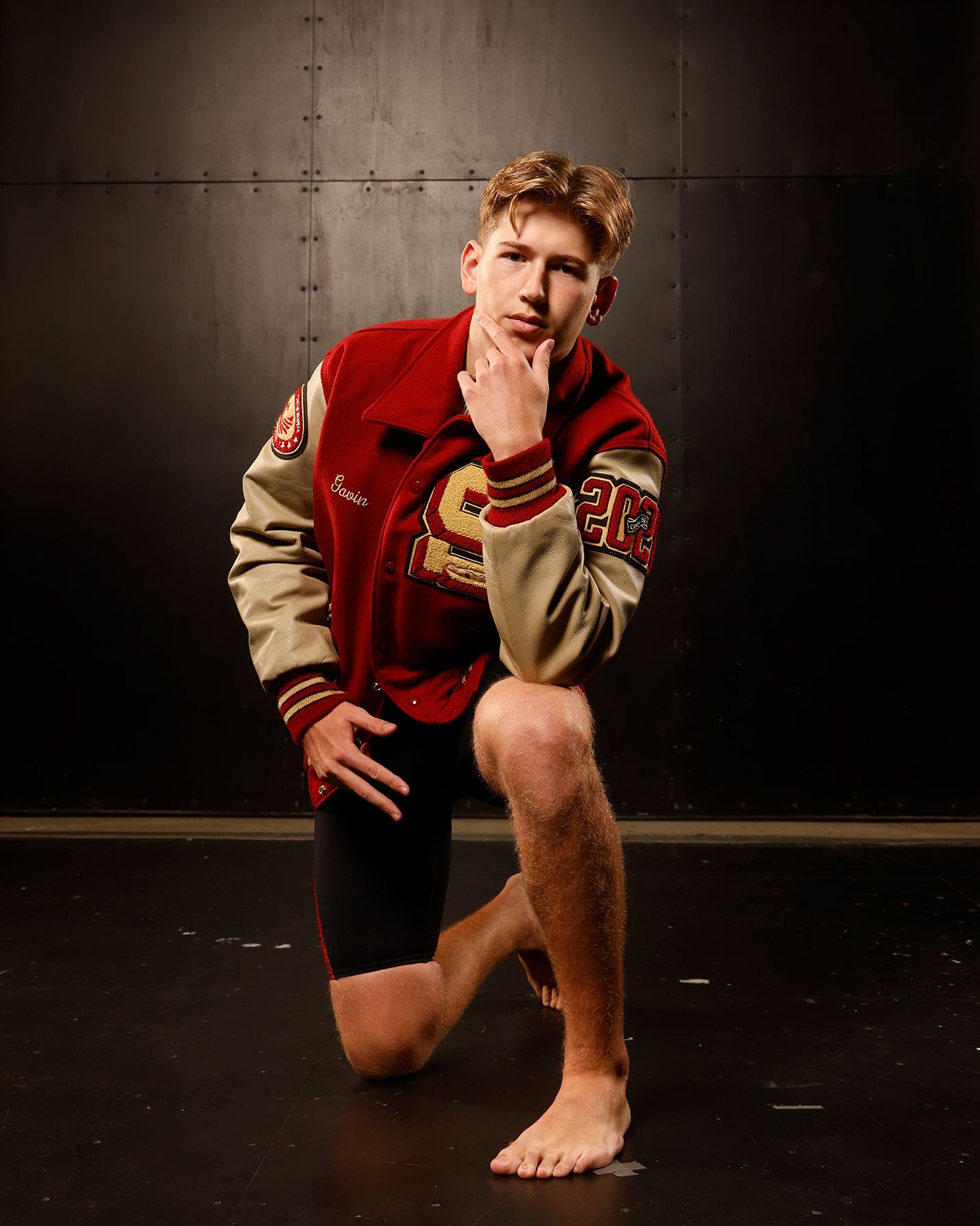 Young athlete kneels in red varsity letterman jacket in dramatic studio portrait.