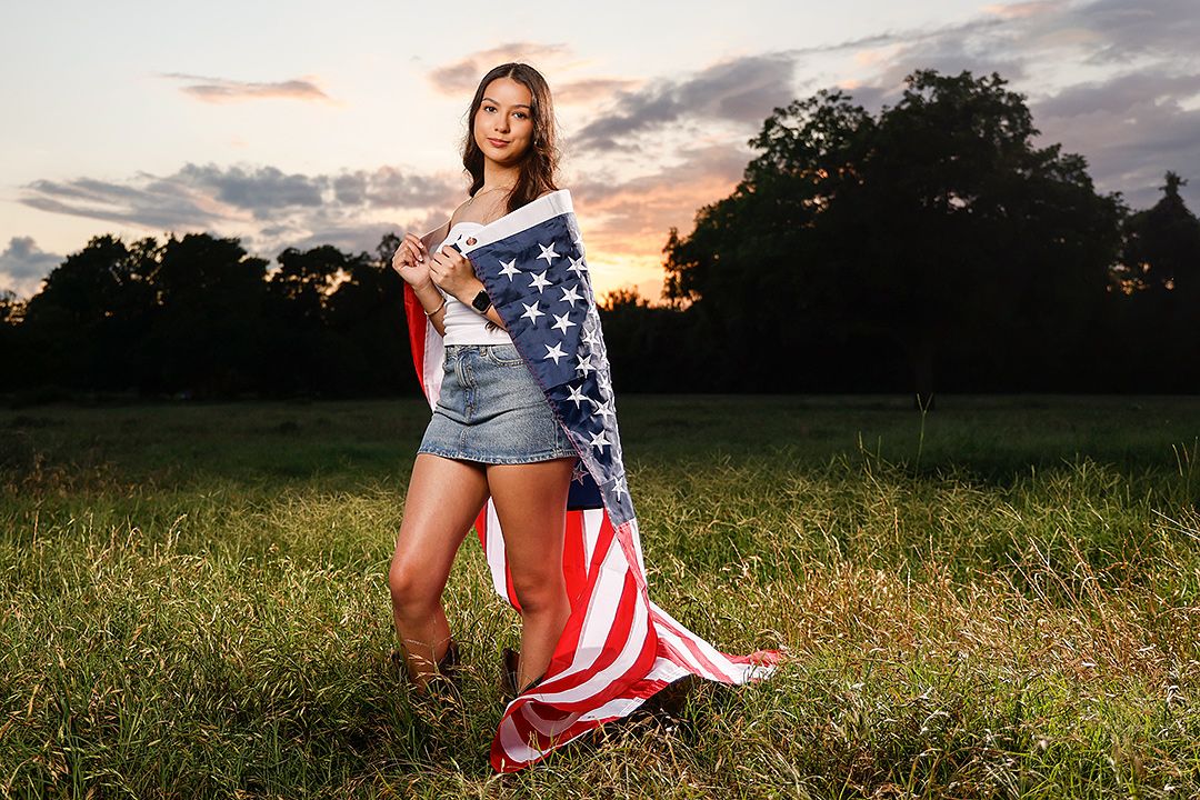 Young woman wrapped in an American flag standing in a green field at sunset with dramatic cloudy sky.