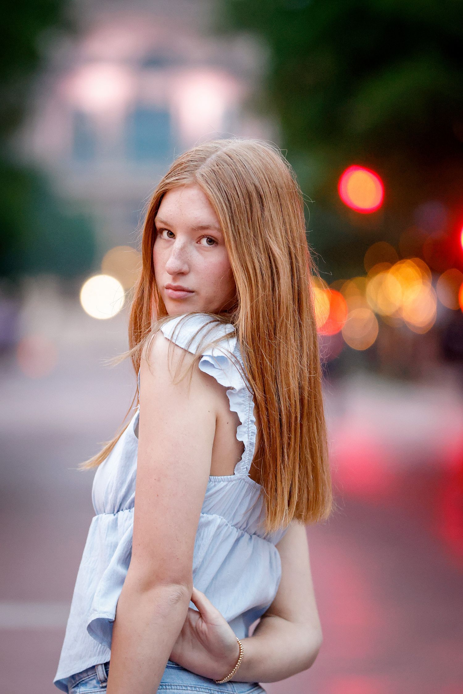 Young woman with long red hair in a light blue top poses on a city street at dusk with bokeh lights.