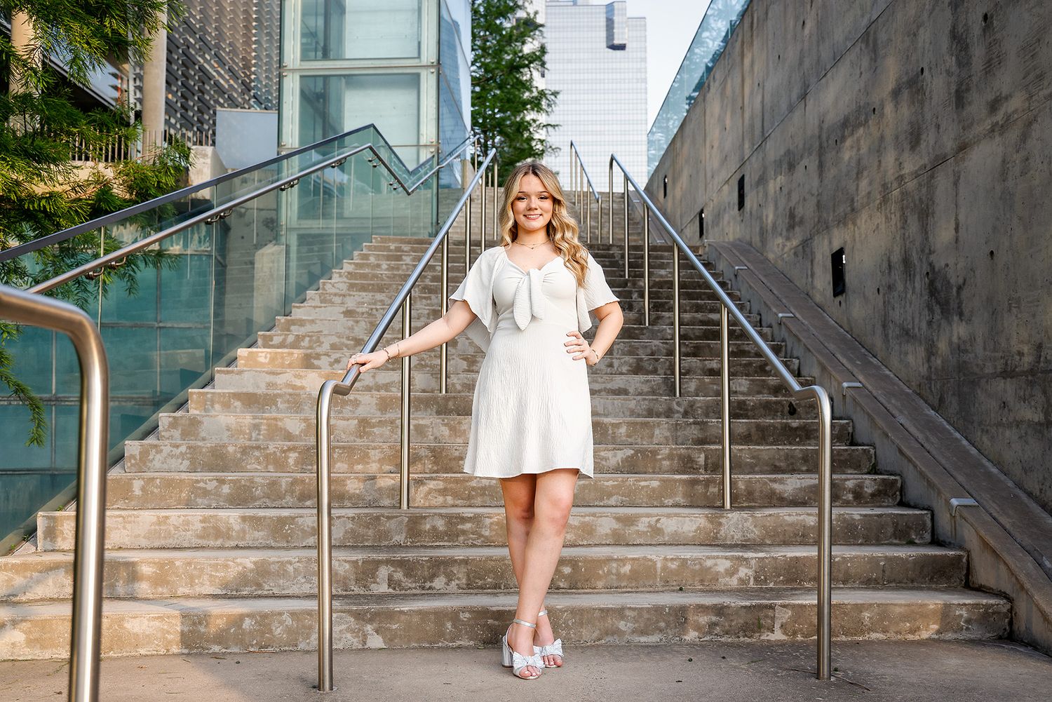 Woman in white dress posing confidently on urban stone steps with glass building backdrop.