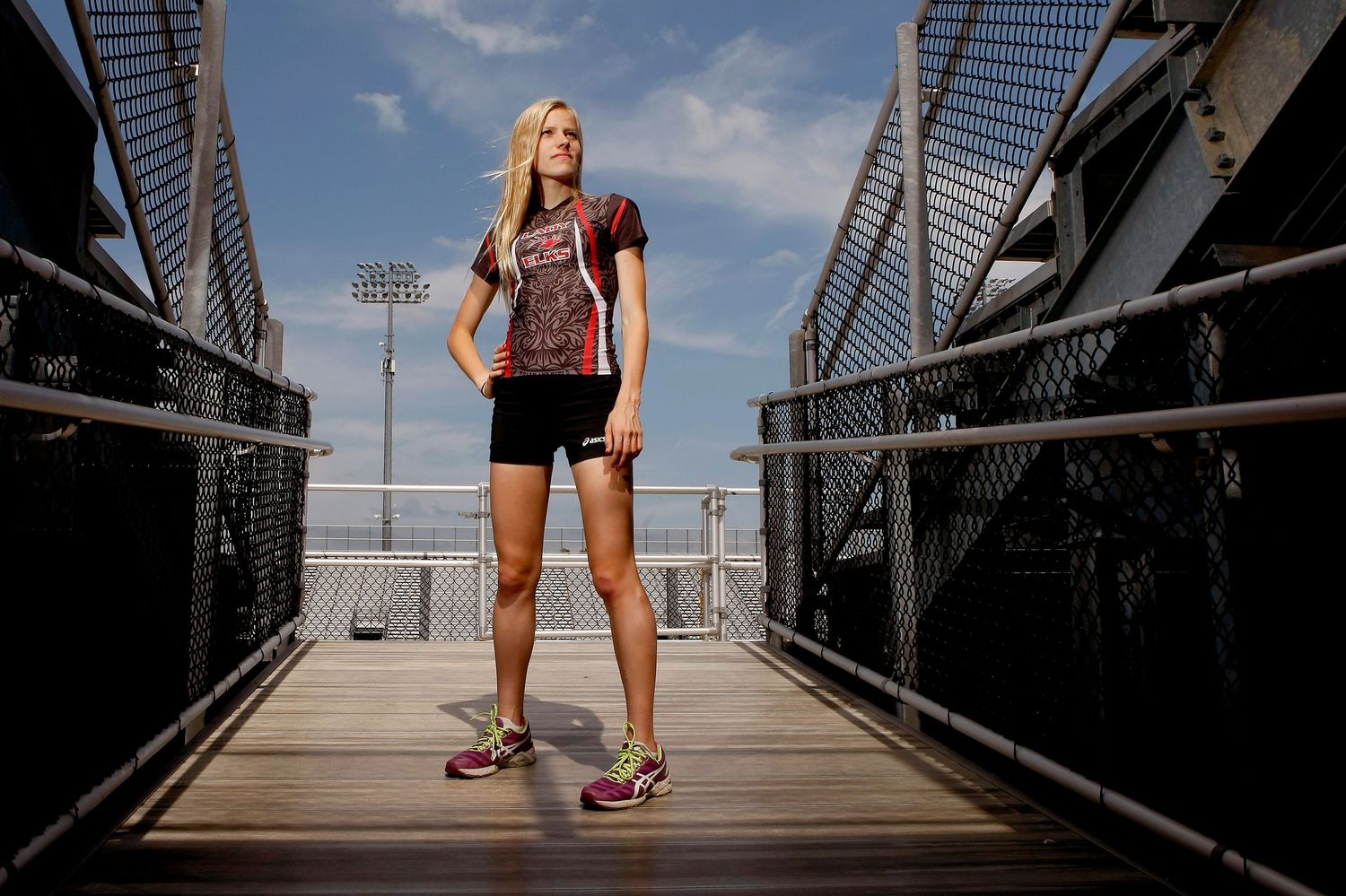 Female athlete in red uniform poses confidently on stadium bridge under bright blue sky.