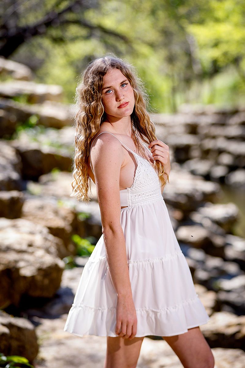 Young woman with curly hair in a white sundress poses on rocky natural stone steps outdoors.