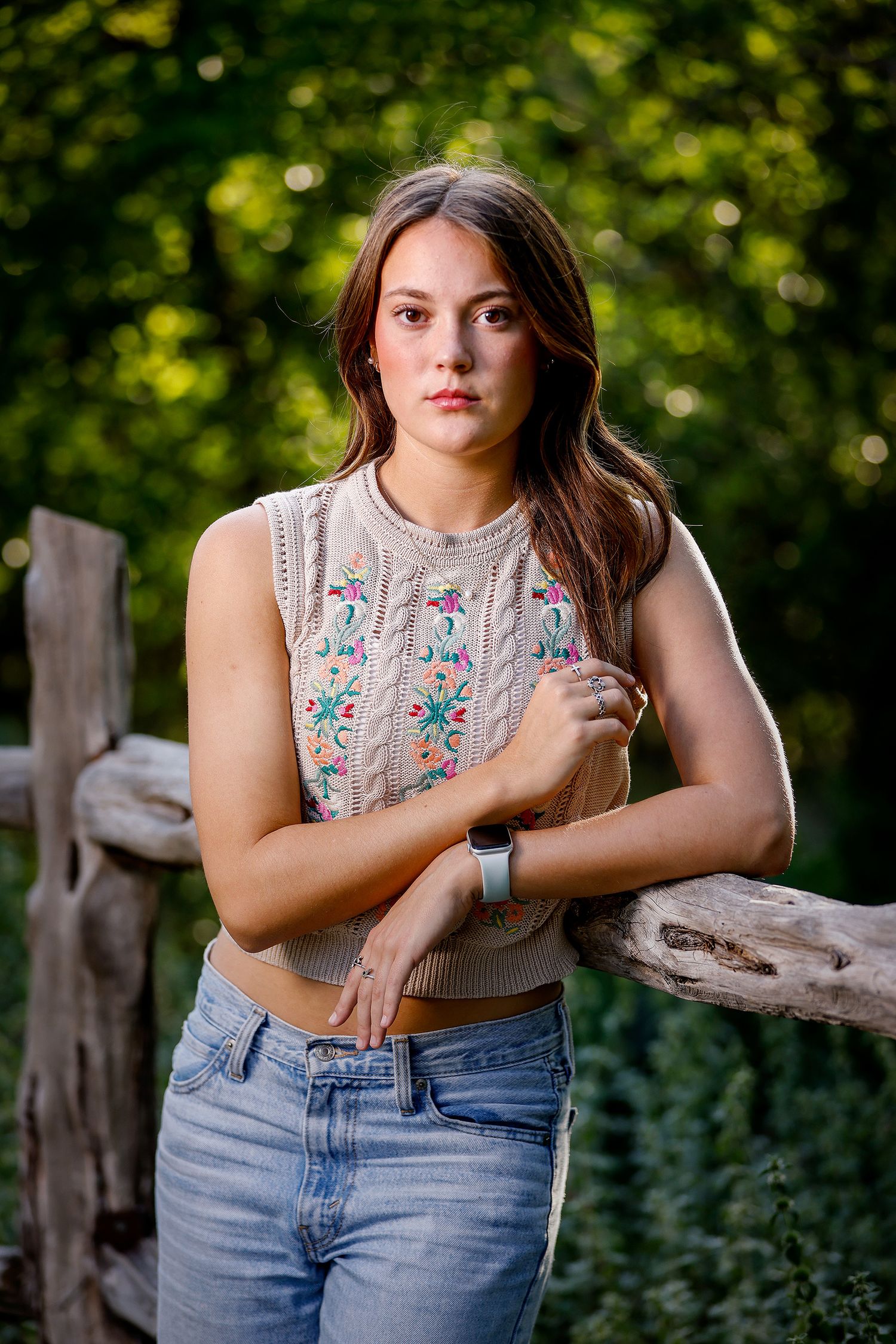 Young woman in floral embroidered crop sweater and jeans leaning on rustic wooden fence outdoors.