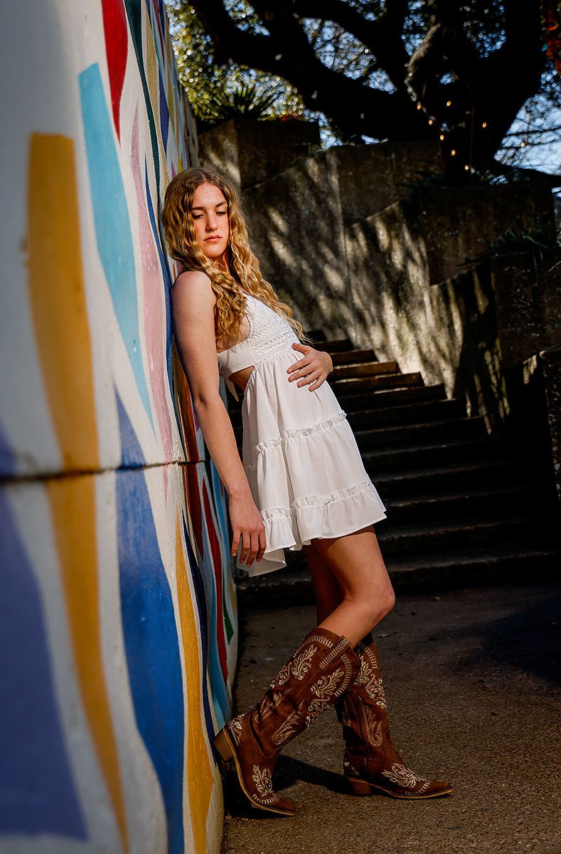Young woman in white dress and cowboy boots leans against a vibrant painted mural wall outdoors.