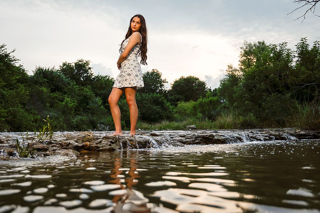 Young woman in floral dress standing on rocks over a serene river with lush green trees in the background.