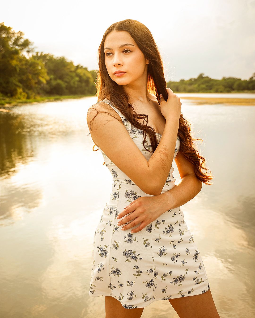 Young woman in floral sundress poses gracefully by a sunlit river surrounded by lush green trees.
