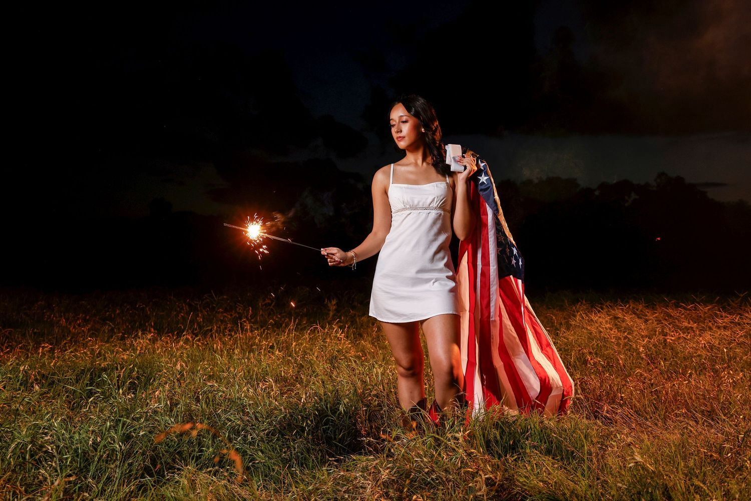 Woman in white dress holding sparkler and American flag in a field at night.