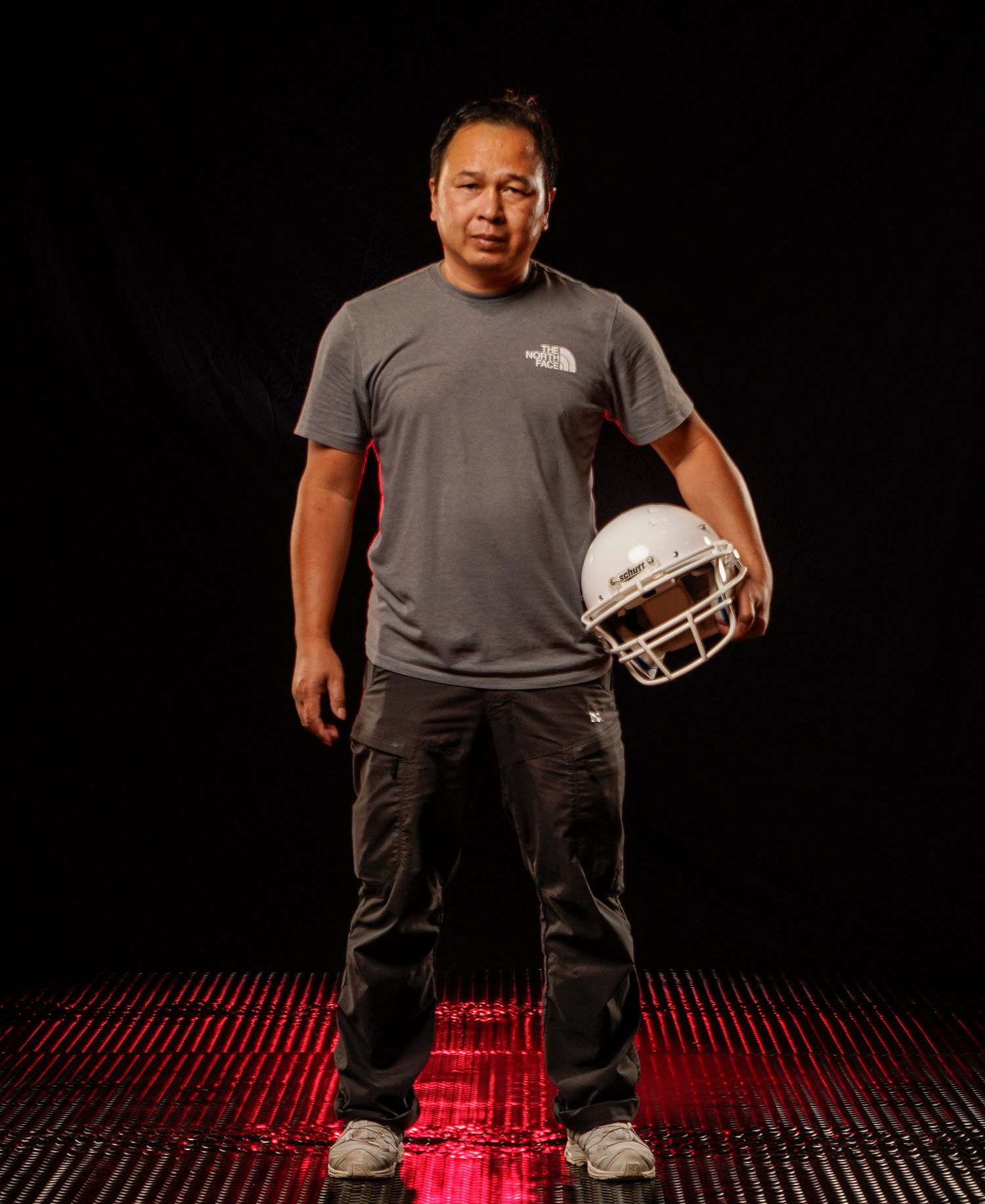 Man in gray North Face shirt holds a white football helmet against a dark background.
