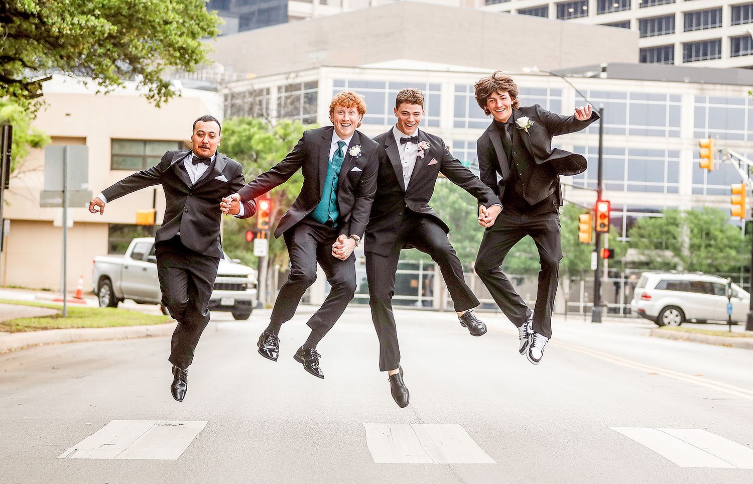 Four groomsmen in black tuxedos joyfully jumping in unison on a city crosswalk during a wedding celebration.