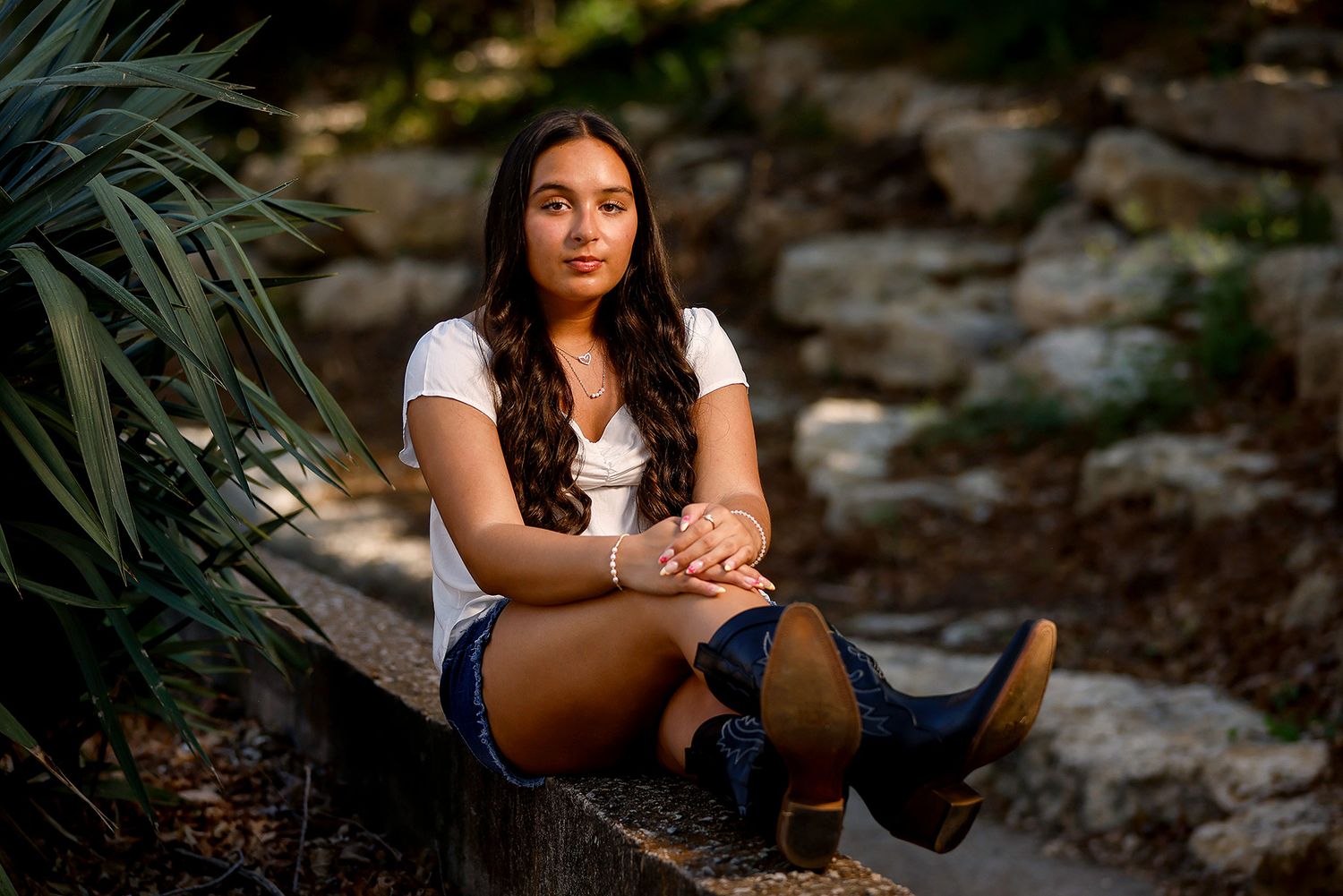 Young woman with long dark hair sits on a wooden bench in a lush garden, wearing a white top and denim shorts.