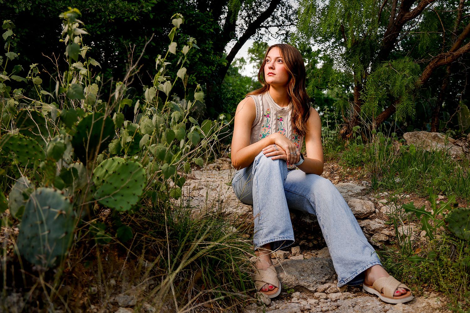 Young woman in flared jeans sitting on rocks surrounded by cacti and greenery, gazing upward outdoors.
