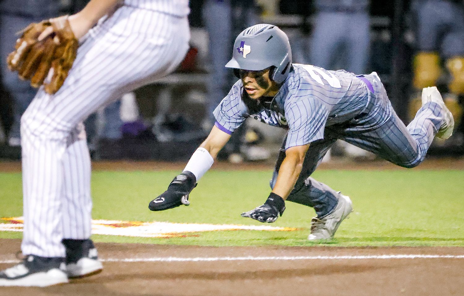 Baseball player diving headfirst into base during a night game while pitcher stands ready on the field.