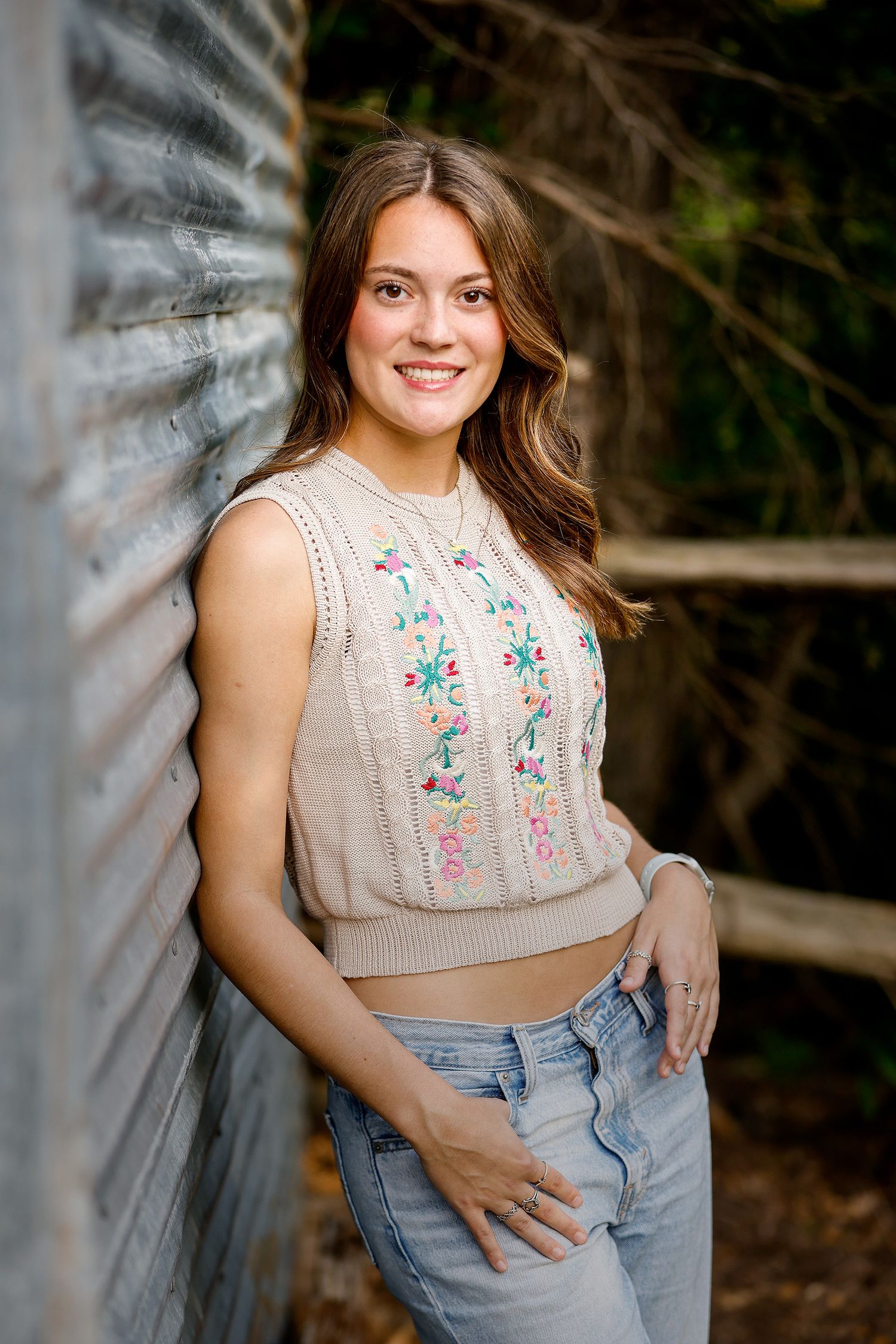 Young woman in floral embroidered knit vest and jeans smiling outdoors against rustic metal wall.