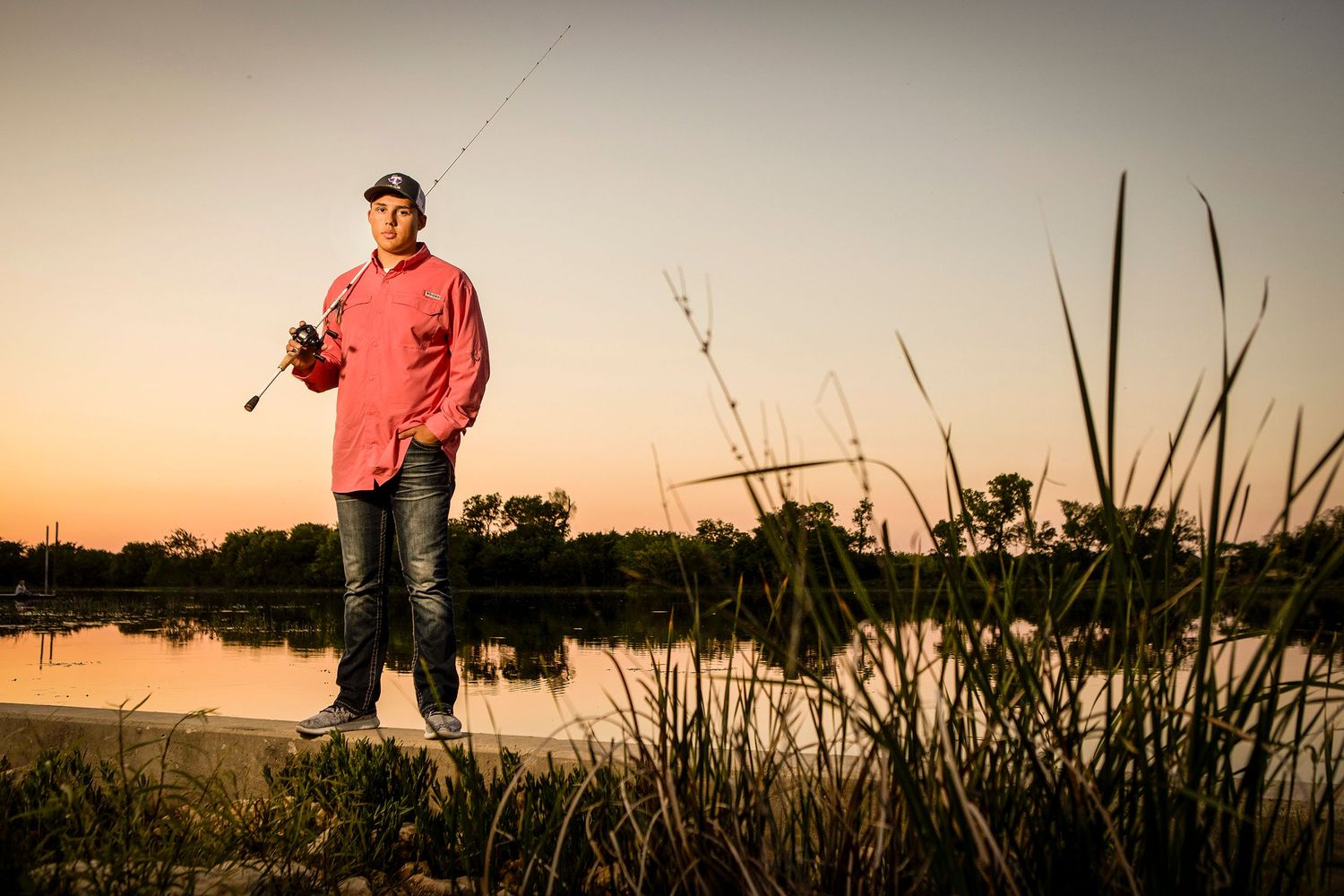 Man in red shirt holds fishing rod while standing by a calm lake at sunset with reeds nearby.