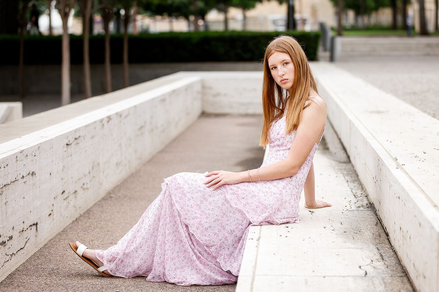 Young woman with long red hair in a floral pink maxi dress sitting gracefully on white stone steps outdoors.