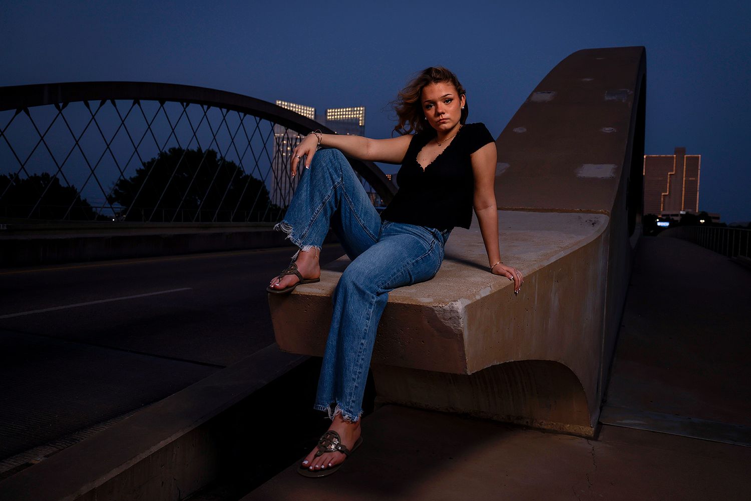 Woman in jeans and black top sitting on concrete bridge ledge at dusk with city lights in background.