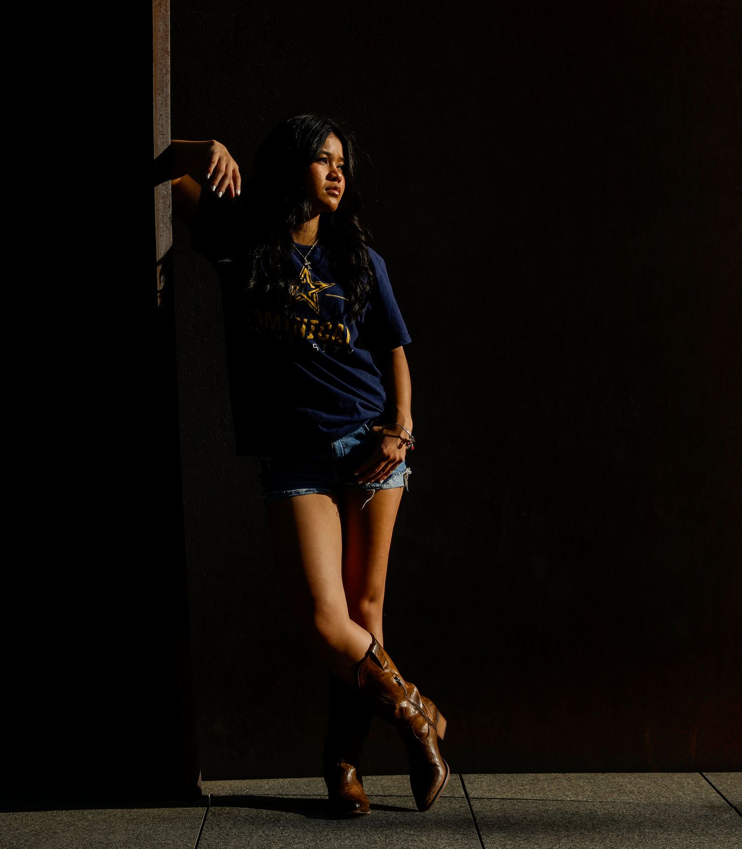 Young woman in blue graphic tee and denim shorts leans against dark wall wearing cowboy boots.