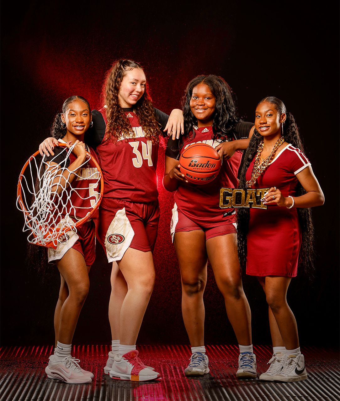 Four female basketball players in red uniforms pose together holding a net, basketball, and championship trophy.