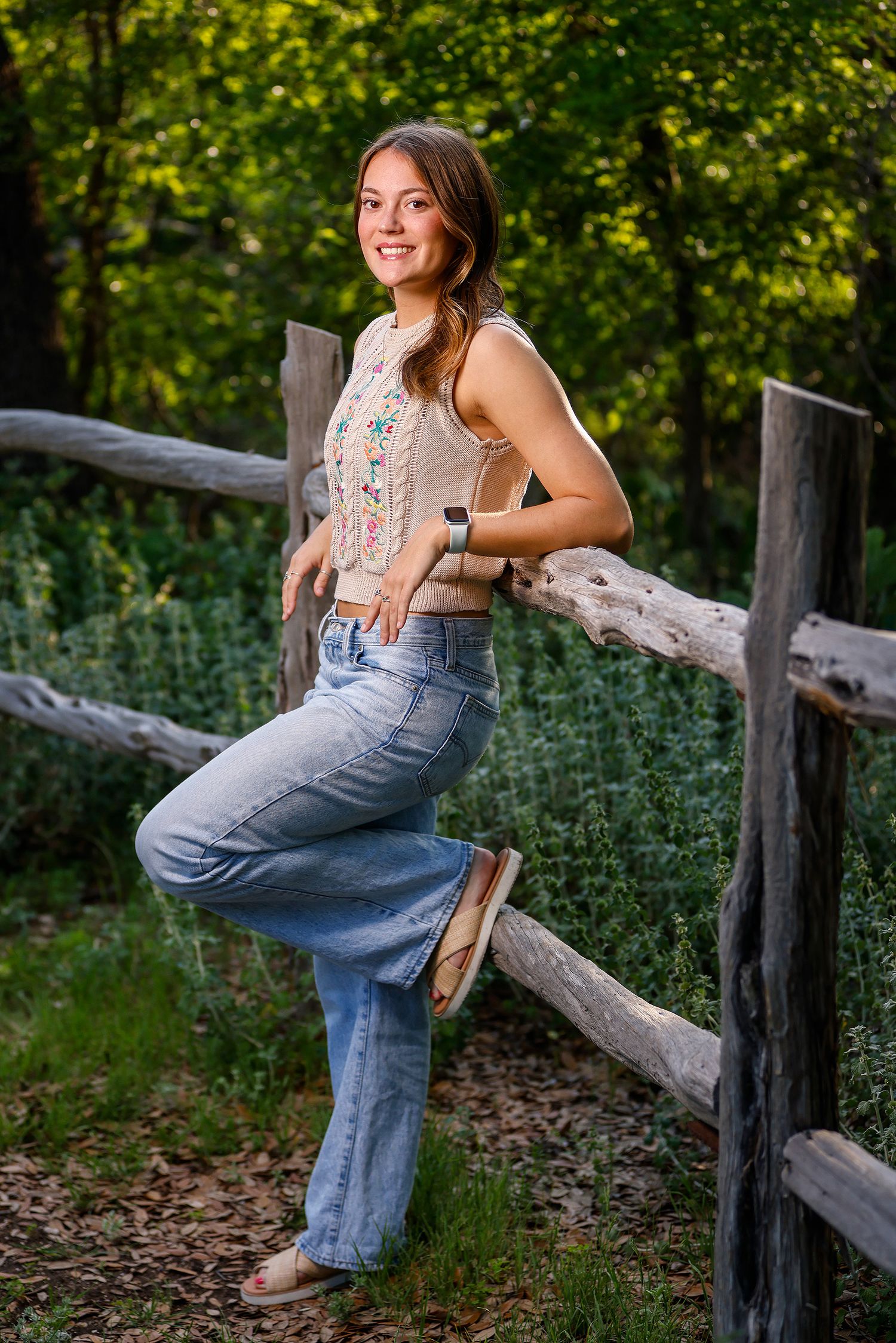 Young woman in floral top and wide-leg jeans leaning on rustic wooden fence surrounded by lush green trees.