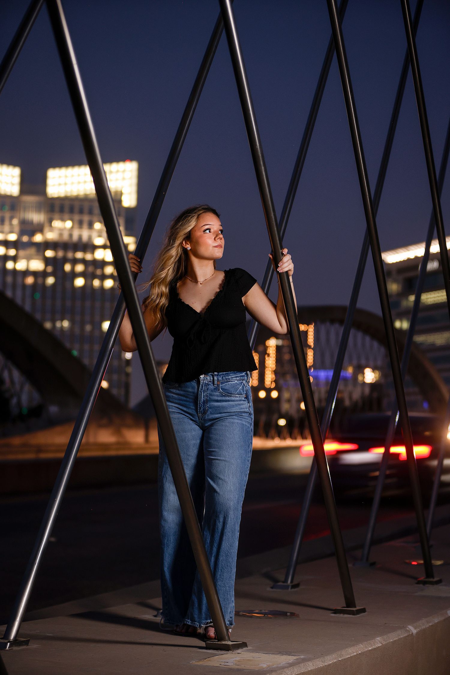 Young woman in black top and jeans poses between metal beams on a bridge with city lights at night.