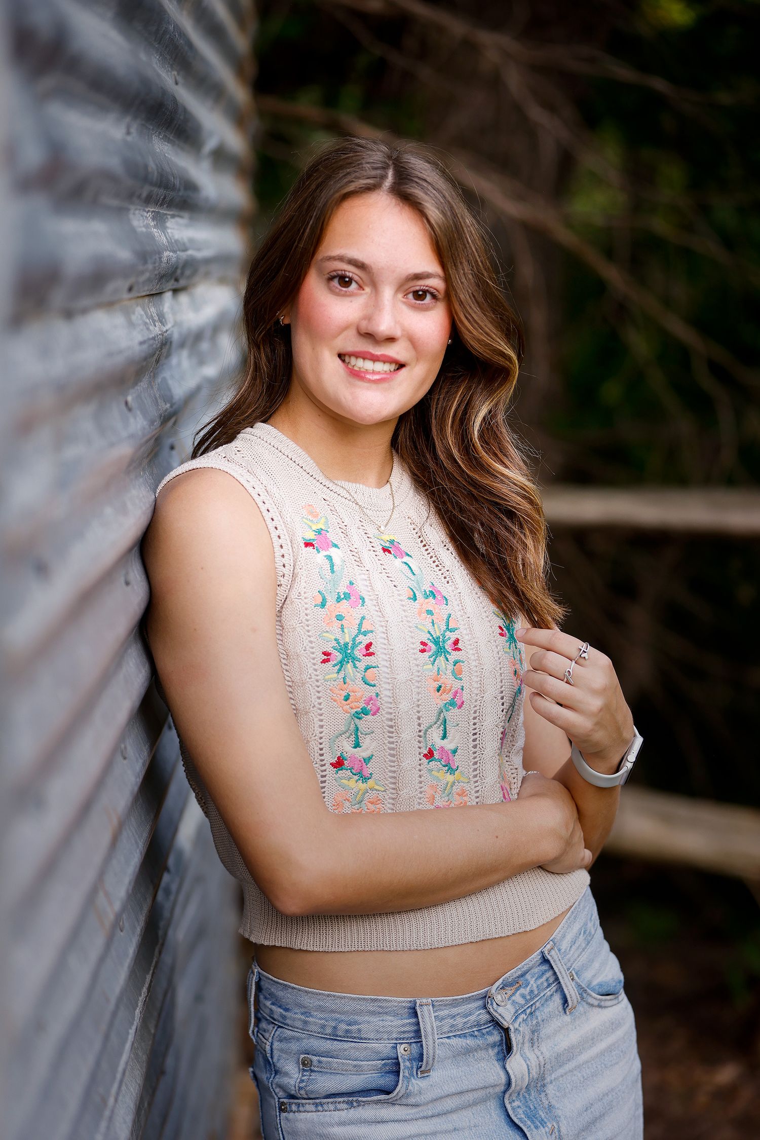 Young woman in floral embroidered vest and jeans smiling outdoors leaning against rustic wooden wall.