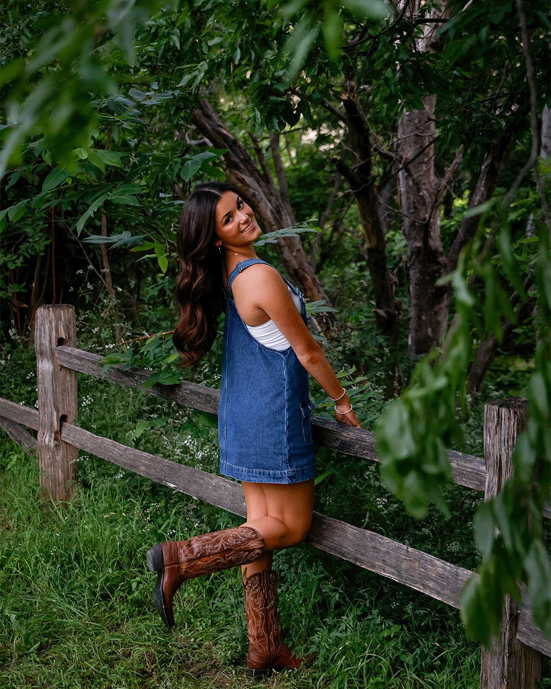 Young woman in denim overalls and cowboy boots poses by a rustic wooden fence surrounded by lush green trees.