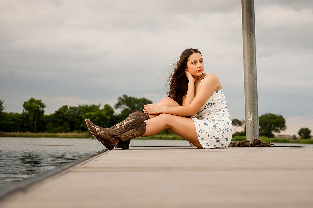 Young woman in white floral dress and cowboy boots sitting on a dock by a lake under cloudy skies.
