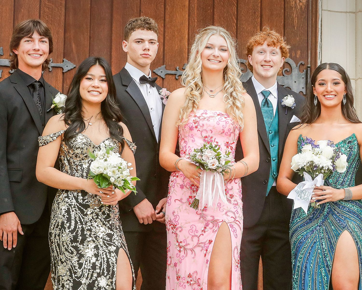 Six teenagers dressed in formal prom attire pose together outdoors, girls holding floral bouquets.