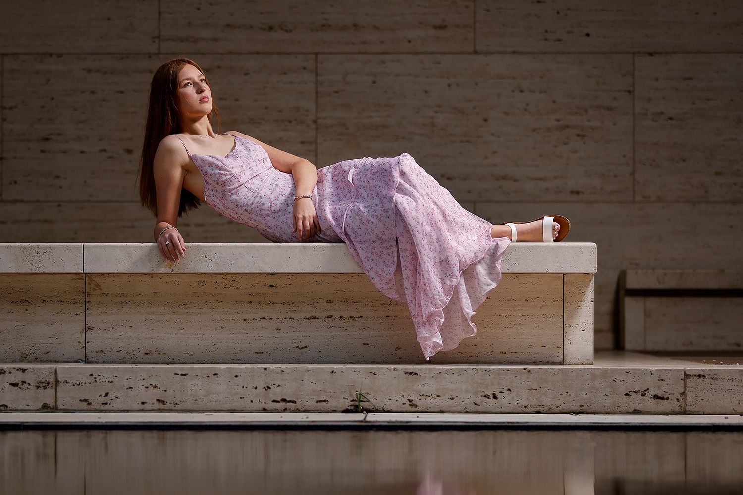 Young woman in a pink floral maxi dress reclining gracefully on a stone bench outdoors.