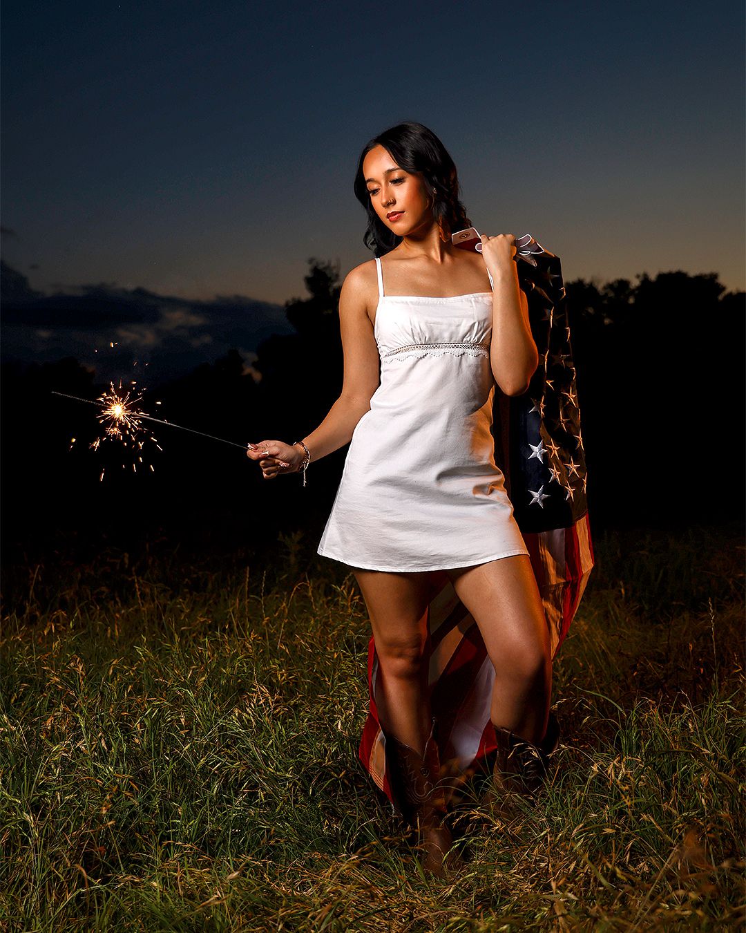 Woman in white dress holding sparkler with American flag at dusk in field wearing cowboy boots.