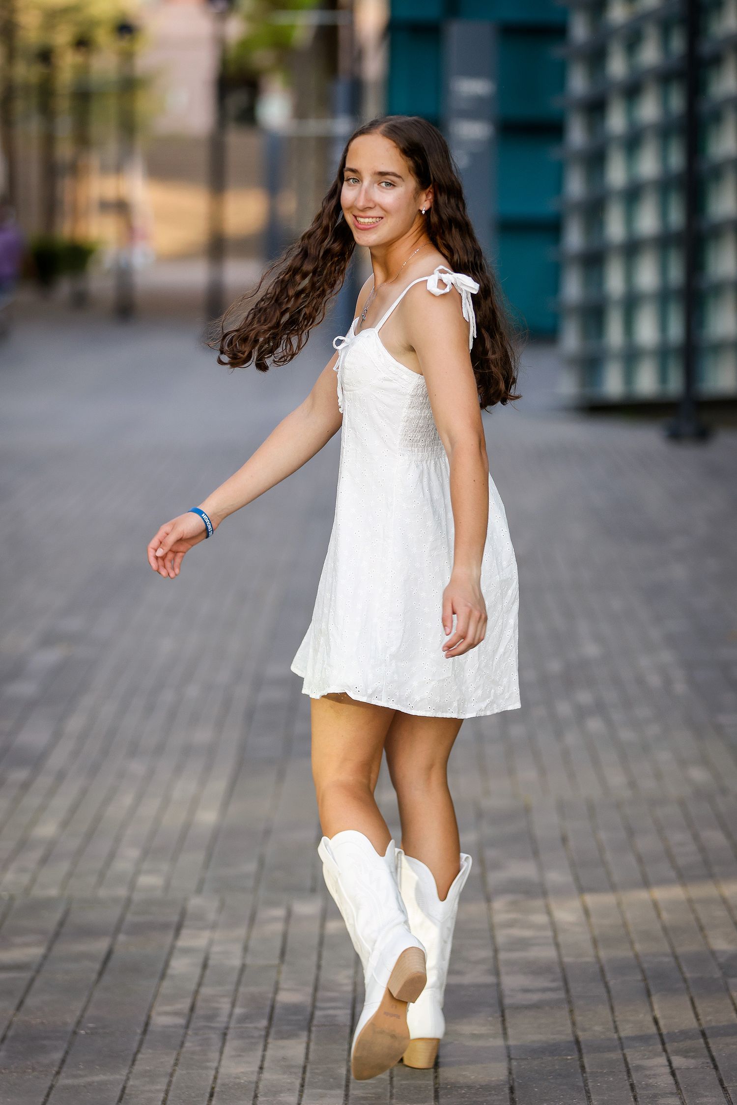 Young woman in white mini dress and white cowboy boots walks on brick pathway, smiling with hair flowing.