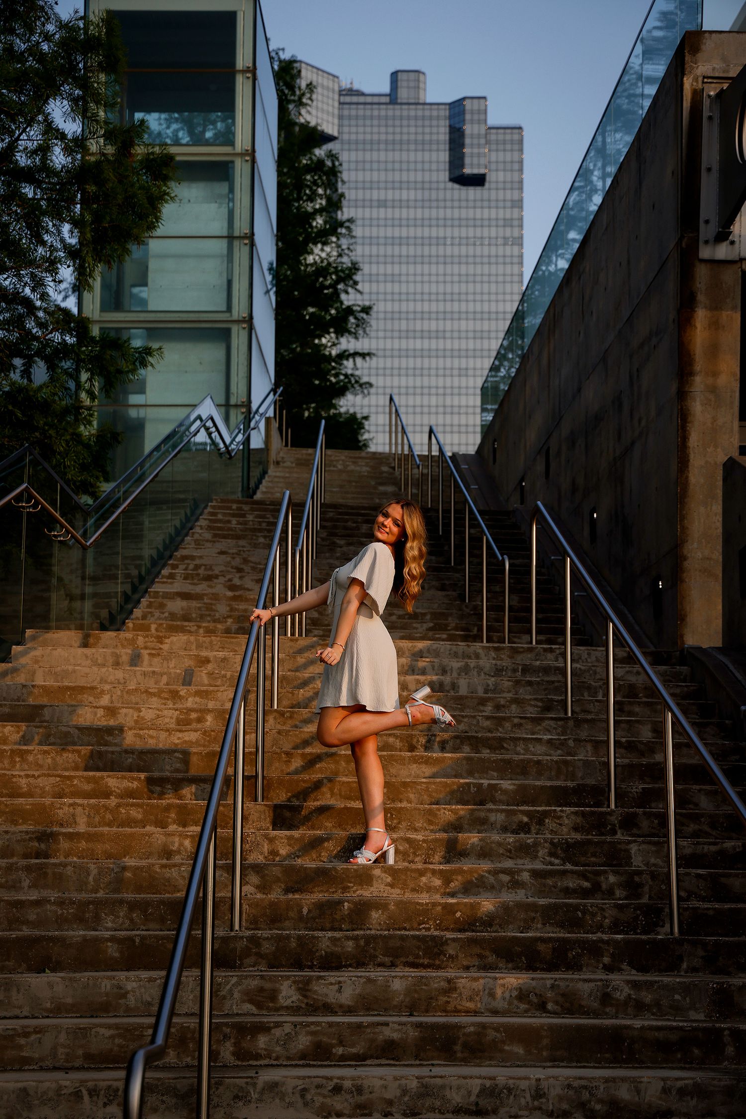 Young woman in white dress posing gracefully on urban stone staircase with modern glass buildings in background.