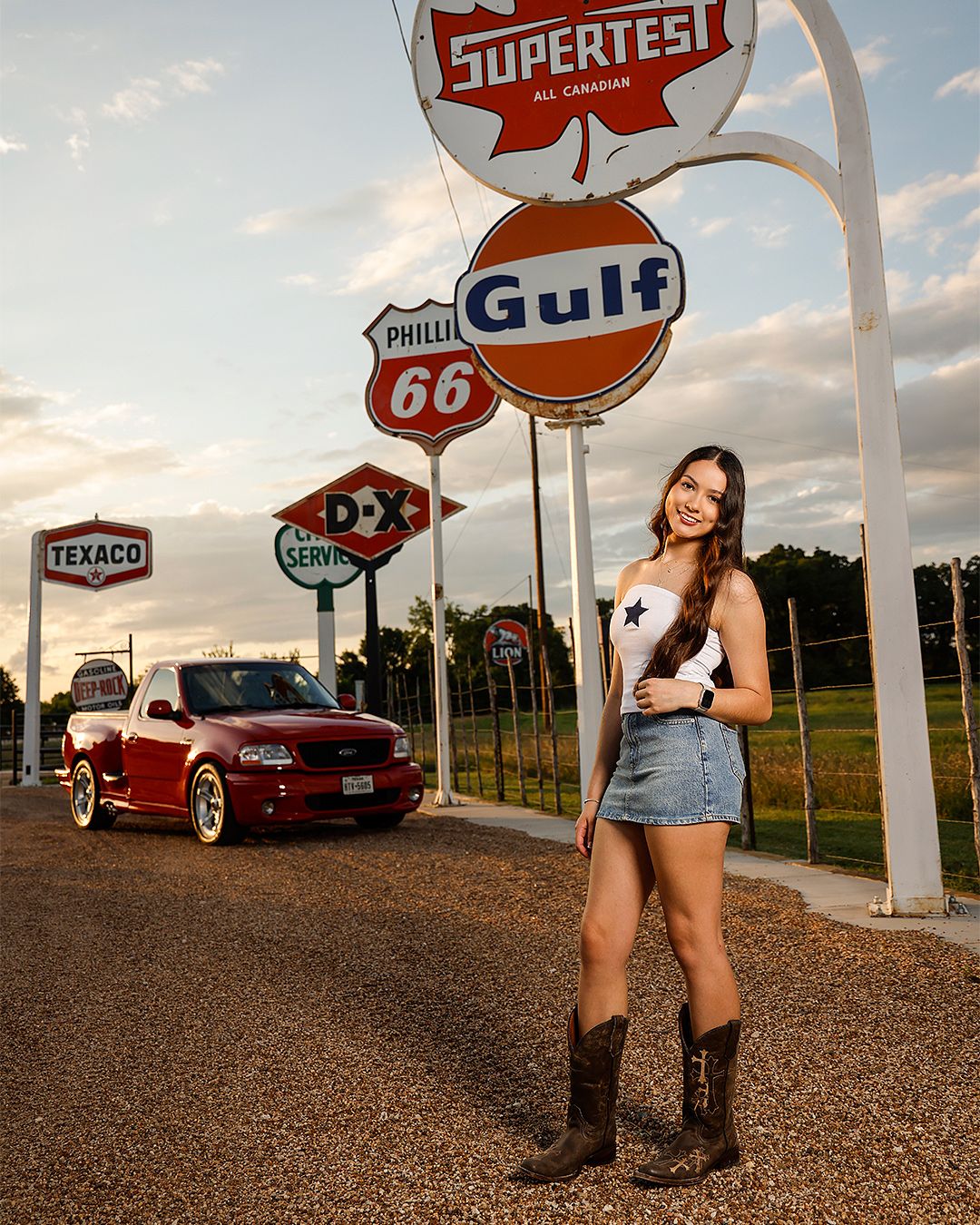 Young woman in denim skirt and cowboy boots poses among vintage gas station signs including Gulf, Texaco, and Phillips 66.