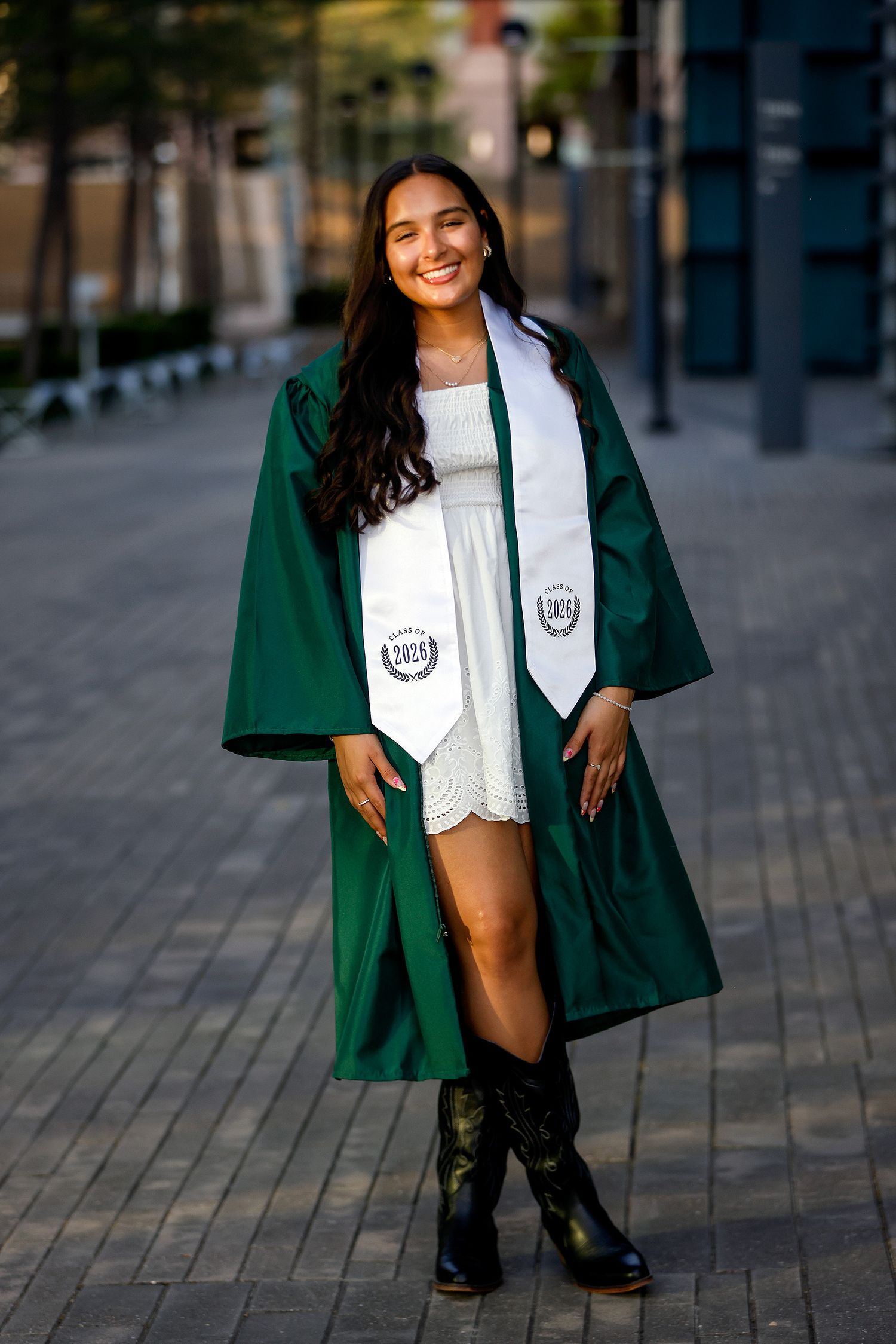 Smiling graduate in green cap and gown with white stole poses outdoors in white dress and black boots.