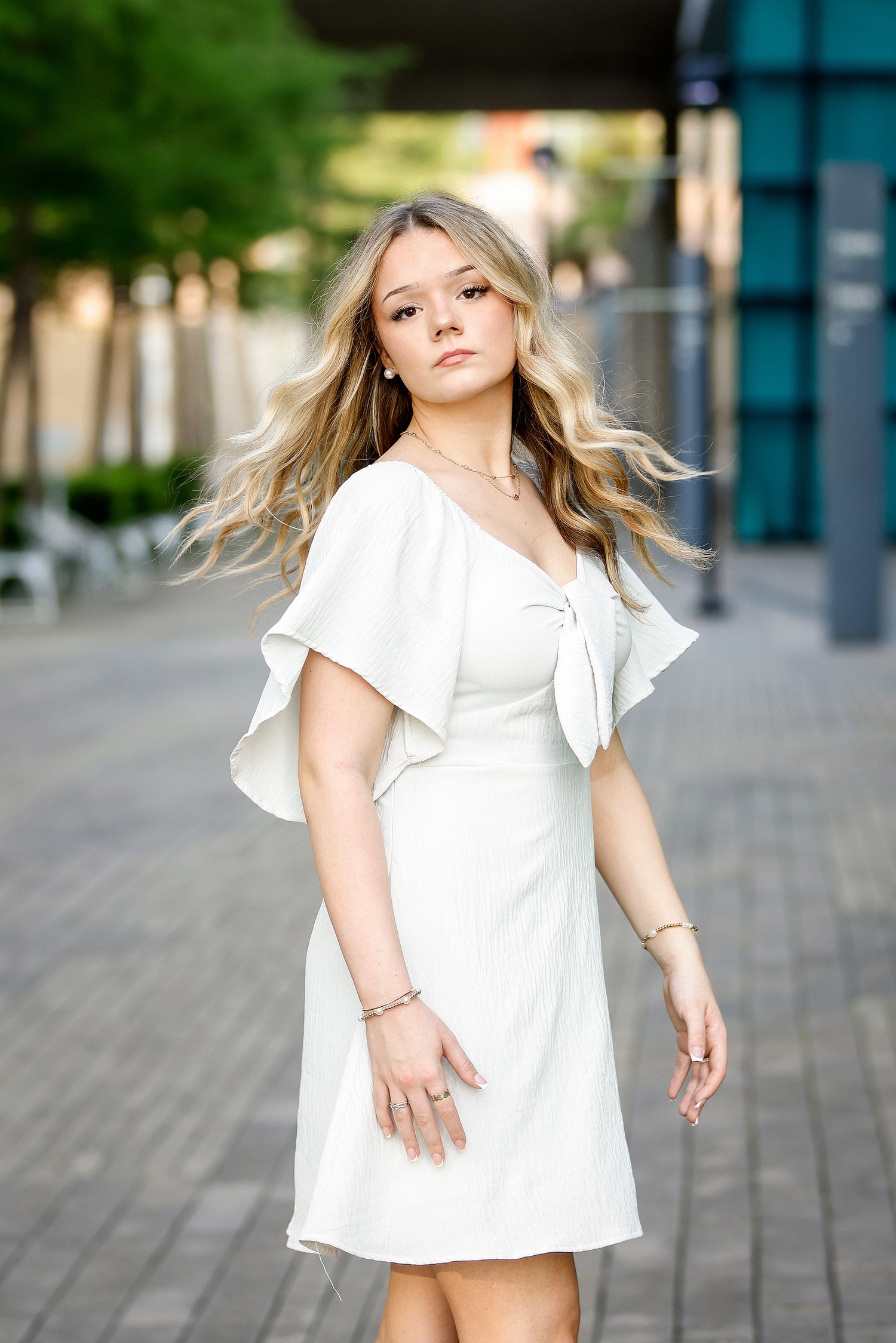 Young blonde woman in white ruffle wrap dress poses outdoors on urban brick walkway with bokeh green background.