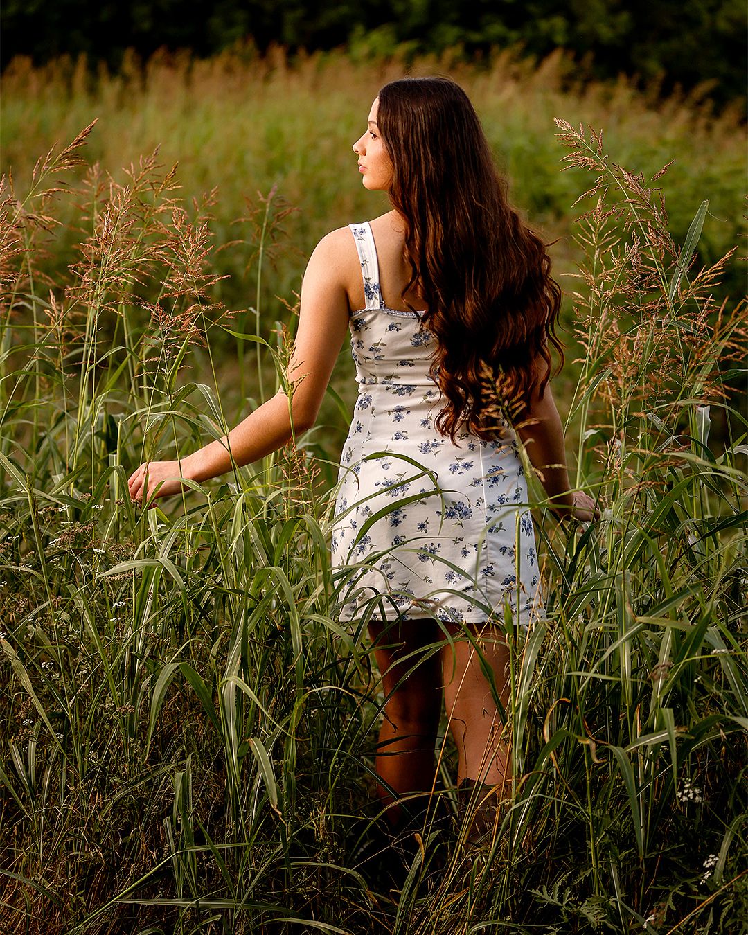 Young woman in floral dress and brown boots standing in tall green grass field, long brunette hair, looking away.