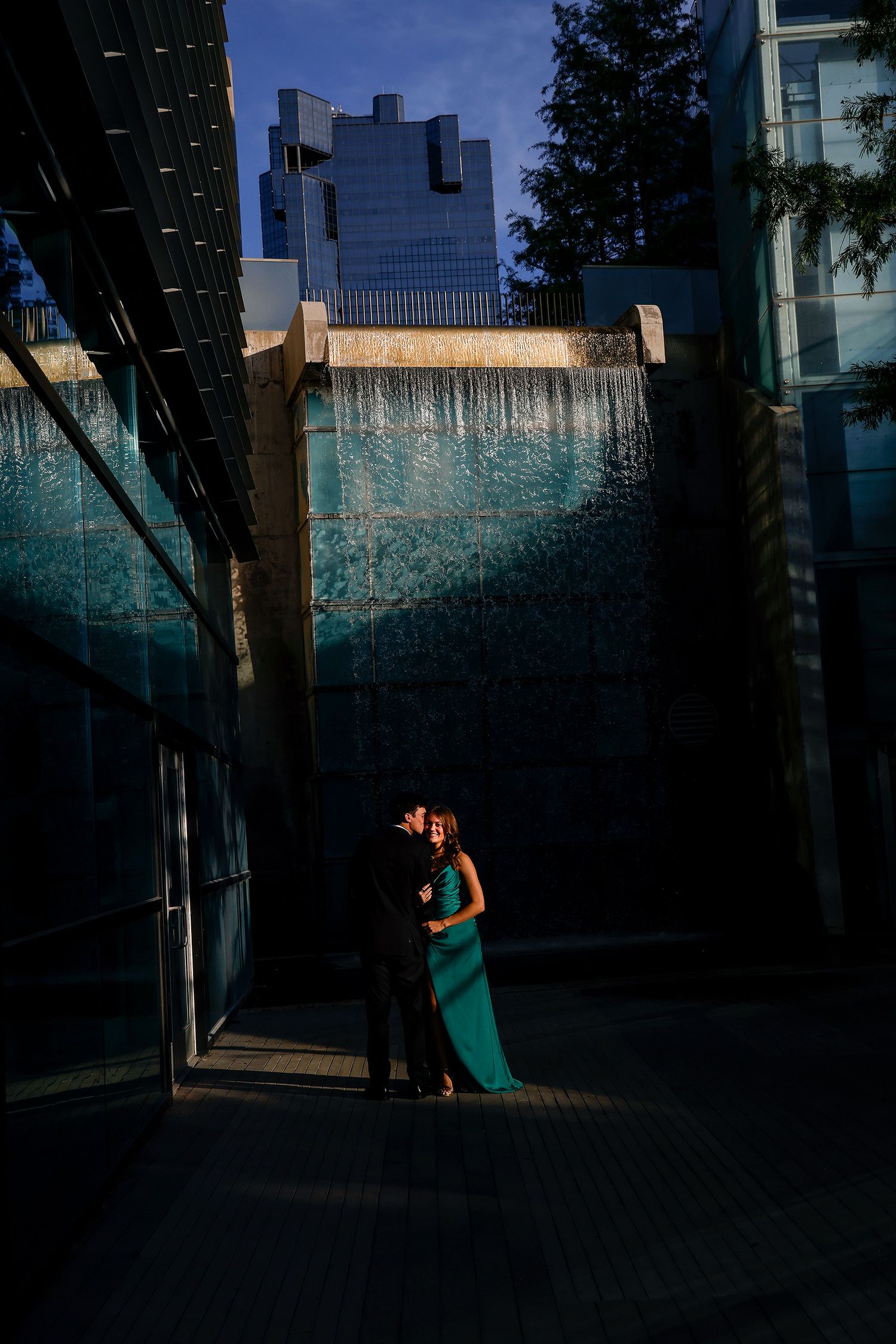 Couple embracing in front of a stunning blue water wall feature at golden hour, woman in elegant teal gown.