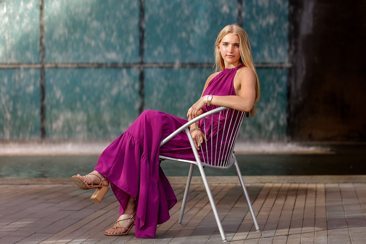 Blonde woman in a flowing magenta jumpsuit seated on a white chair near a fountain outdoors.