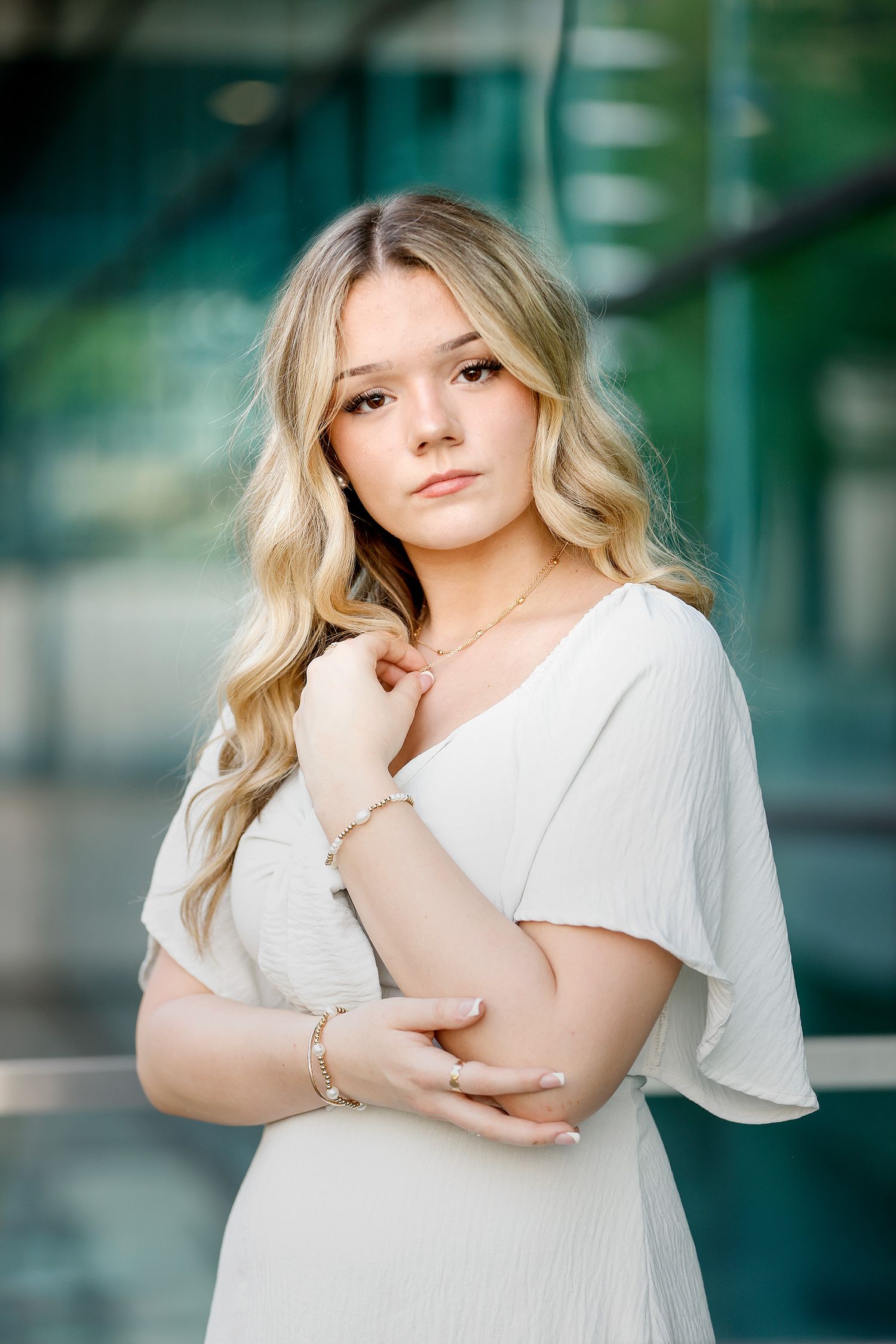 Young blonde woman in white dress posing elegantly with delicate jewelry in a modern glass building.