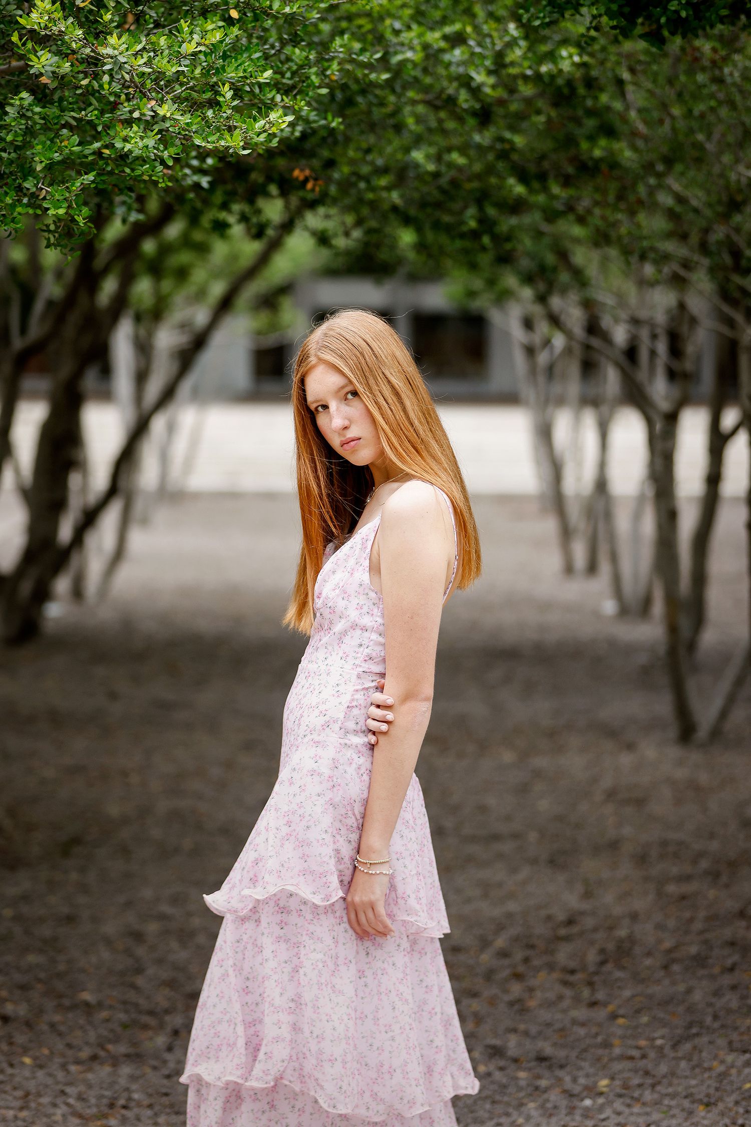 A young woman with long red hair poses in a pink floral tiered dress among green trees outdoors.