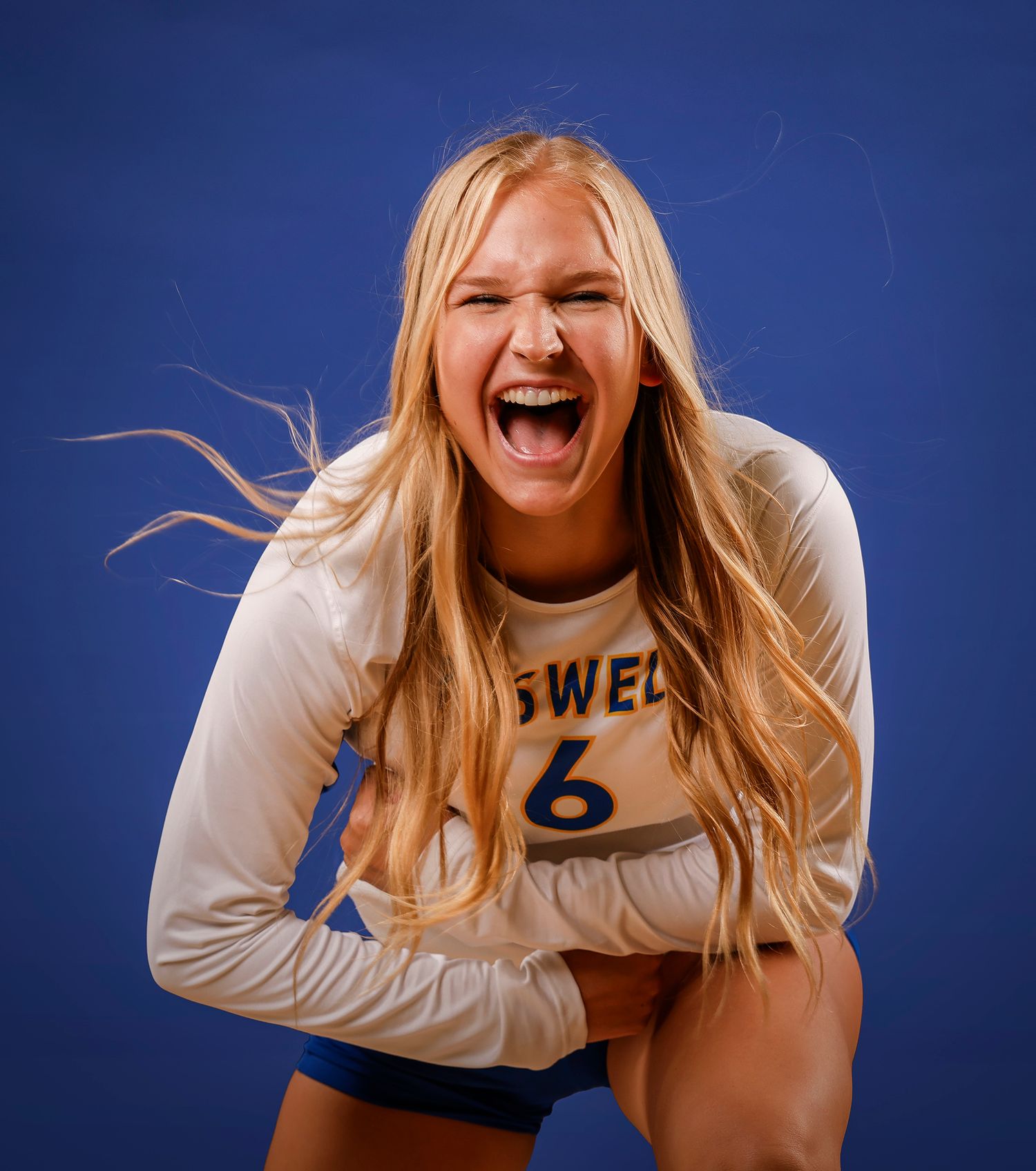 Energetic blonde female volleyball player #6 in white jersey poses with mouth open for team photo.