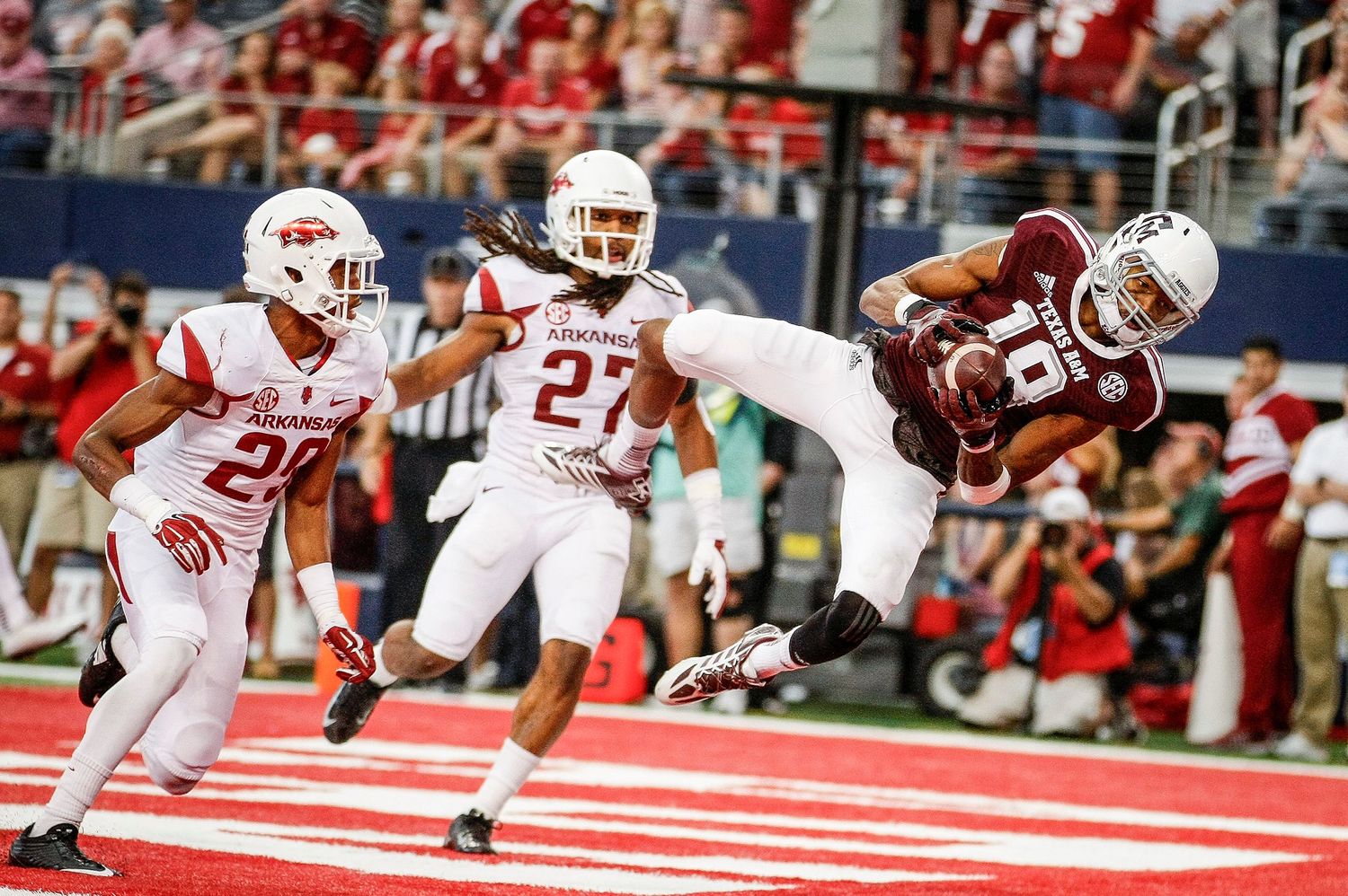 Football player in maroon uniform makes an acrobatic catch while defenders in white pursue during a game.