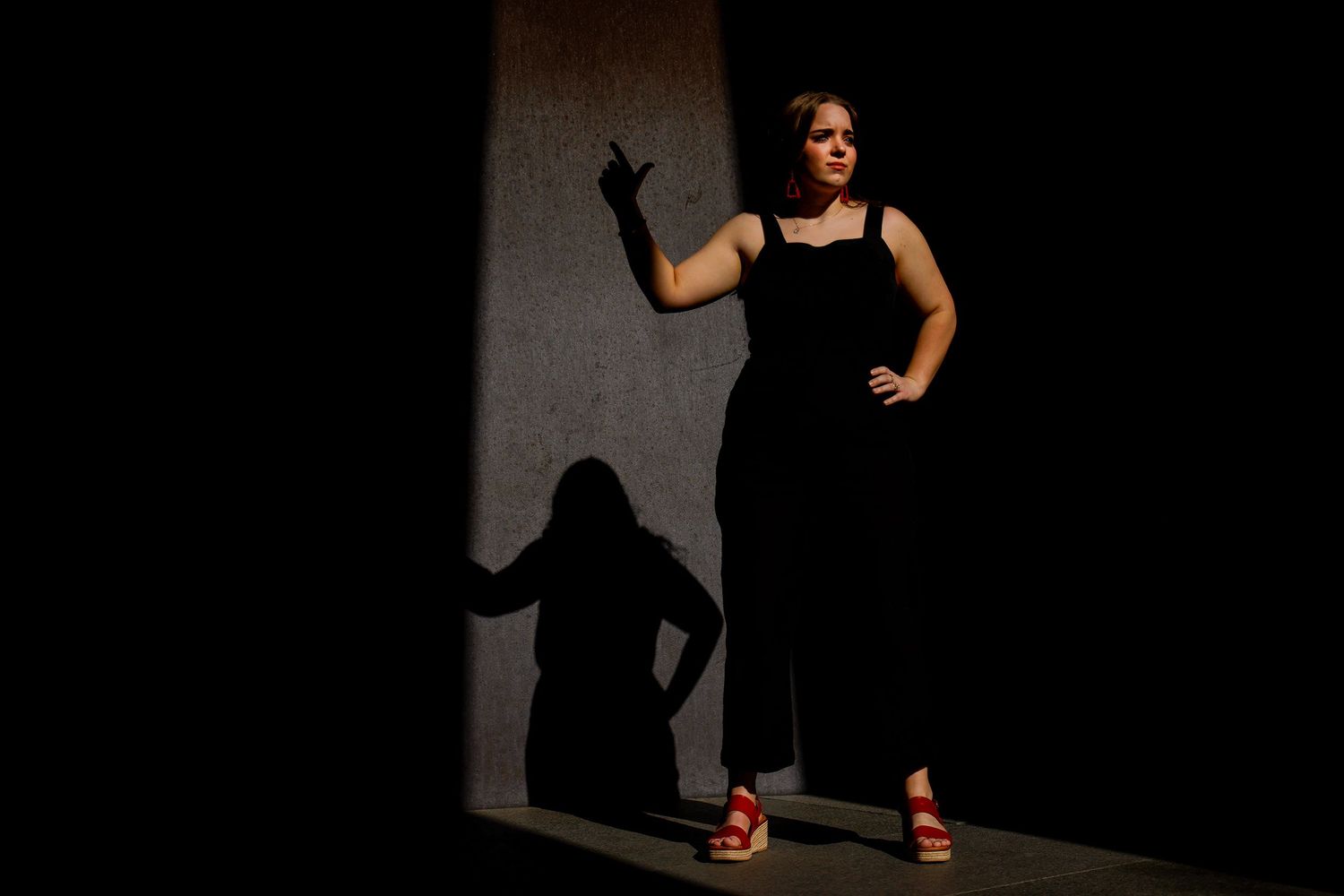 Woman in black dress stands in dramatic light casting a strong shadow on a concrete wall.