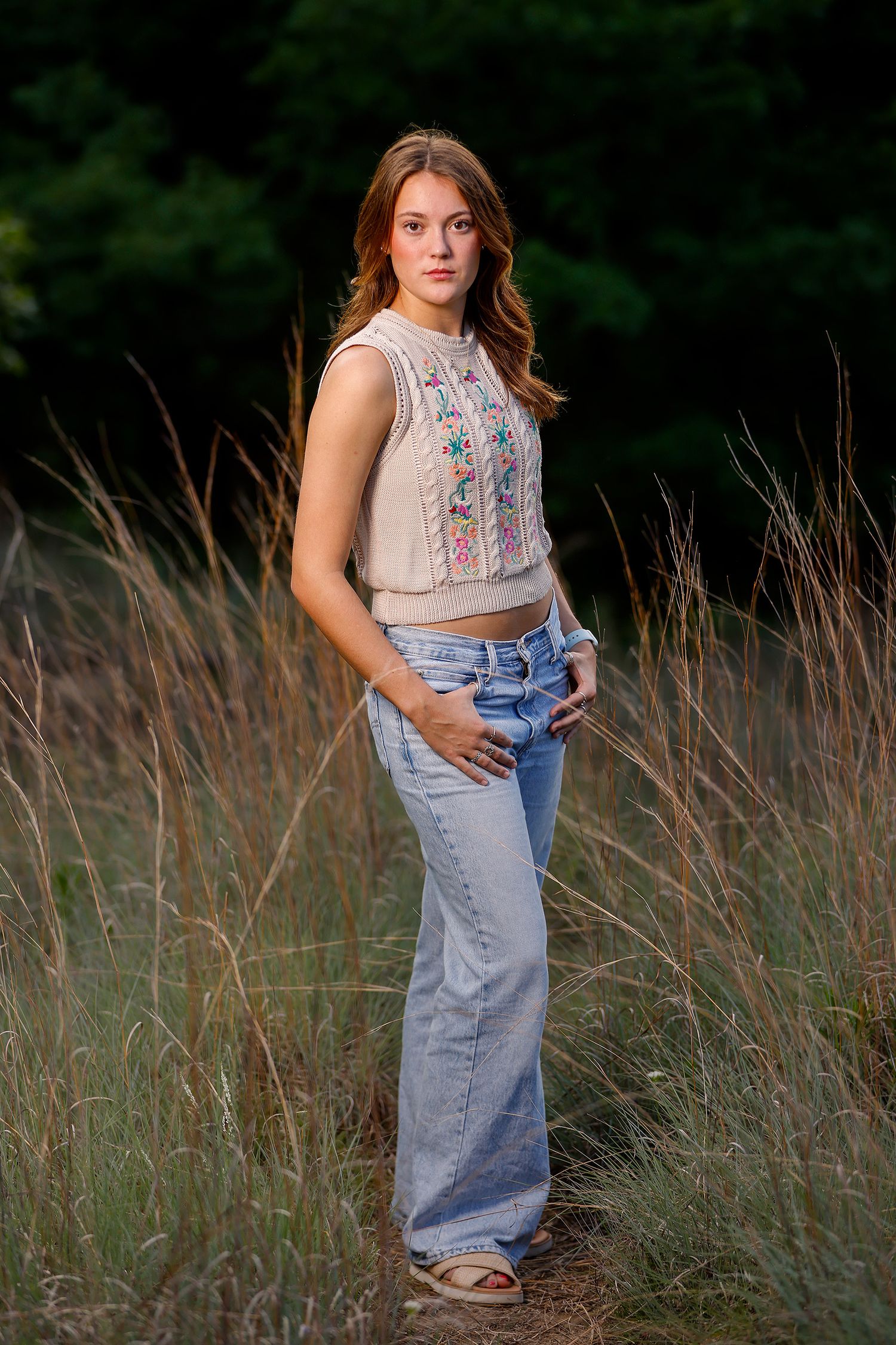 Young woman in floral crop top and wide-leg jeans standing in tall grass field with dark forest background.
