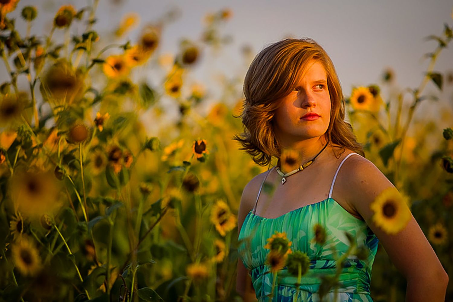 Young woman in green dress standing among blooming sunflowers in golden hour light.