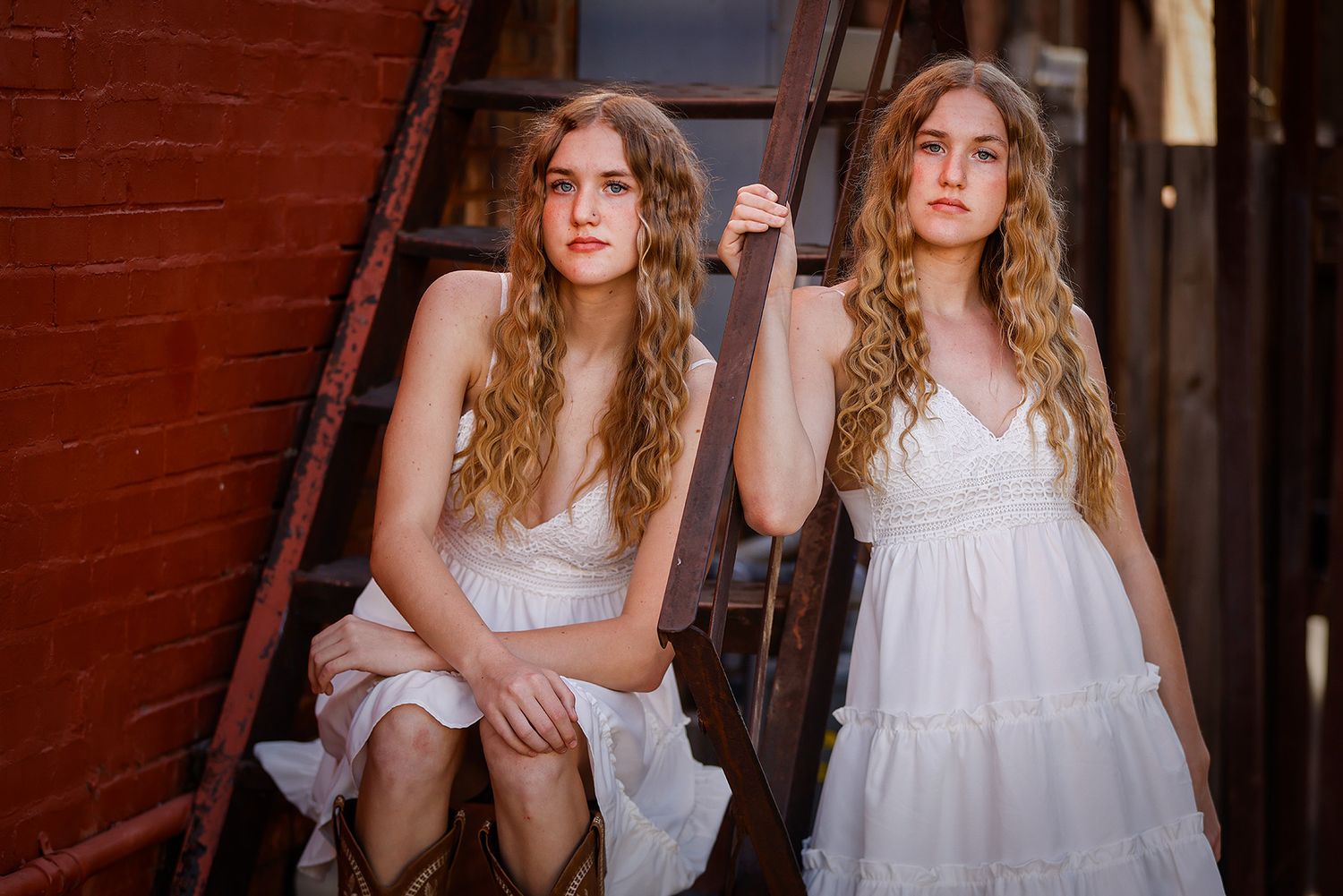 Two young women with curly hair in white dresses pose on a rustic red metal fire escape stairway.