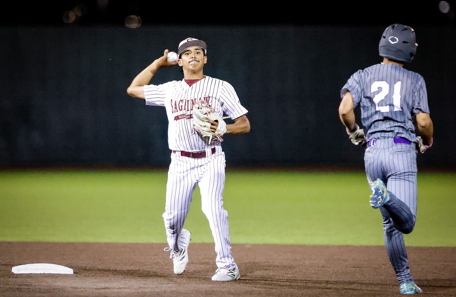 Baseball infielder in maroon uniform throws to first base as opponent in number 21 jersey runs past during night game.