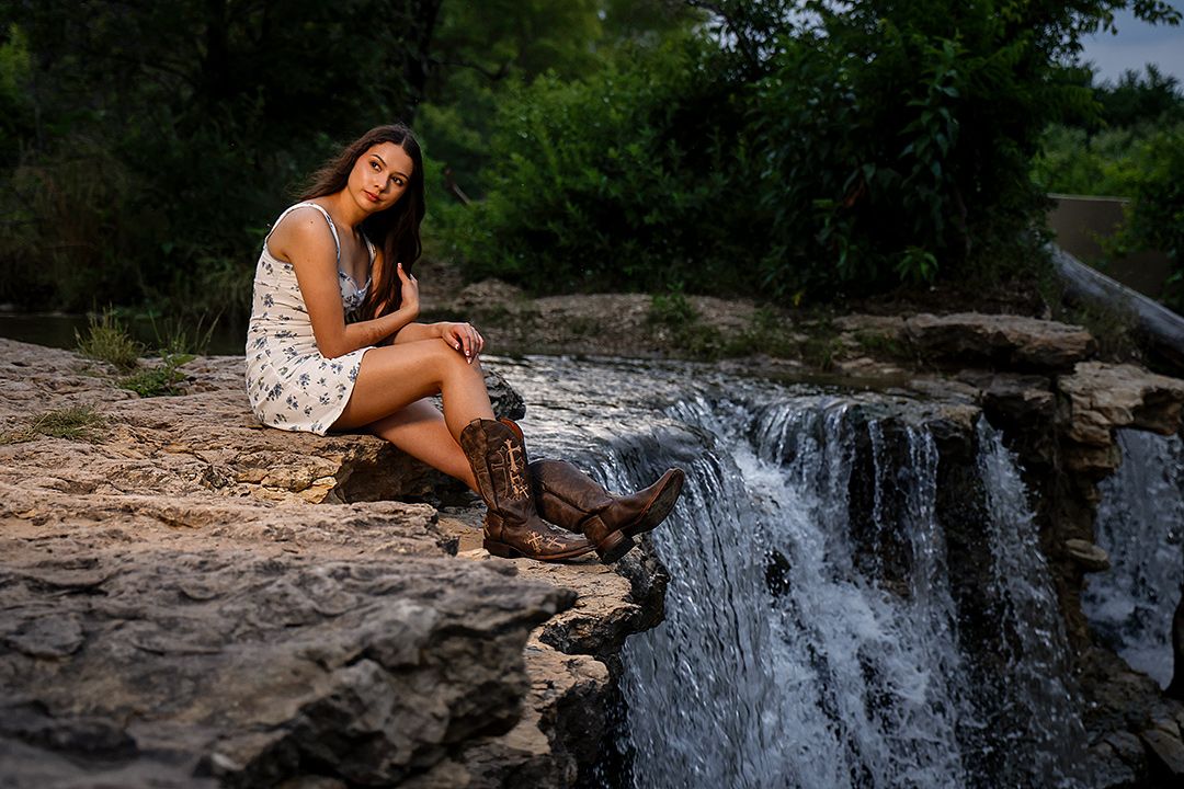 Young woman in white dress and cowboy boots sitting on rocks beside a cascading waterfall surrounded by lush greenery.