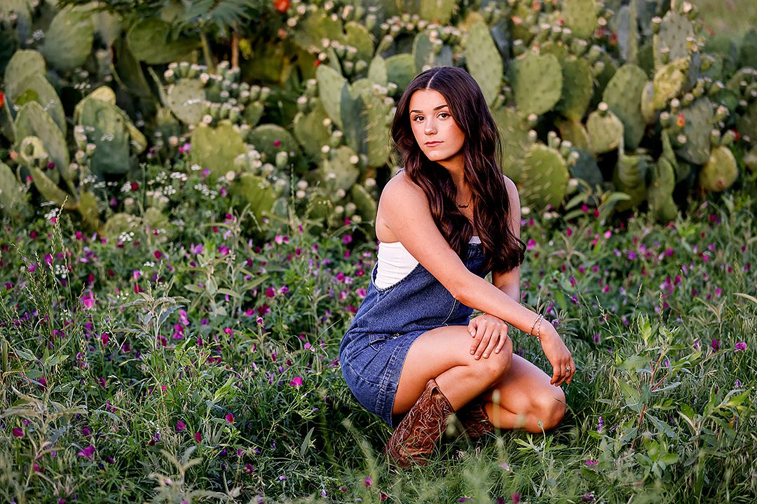 Young woman in white top and blue skirt kneeling among wildflowers and prickly pear cacti in golden hour light.