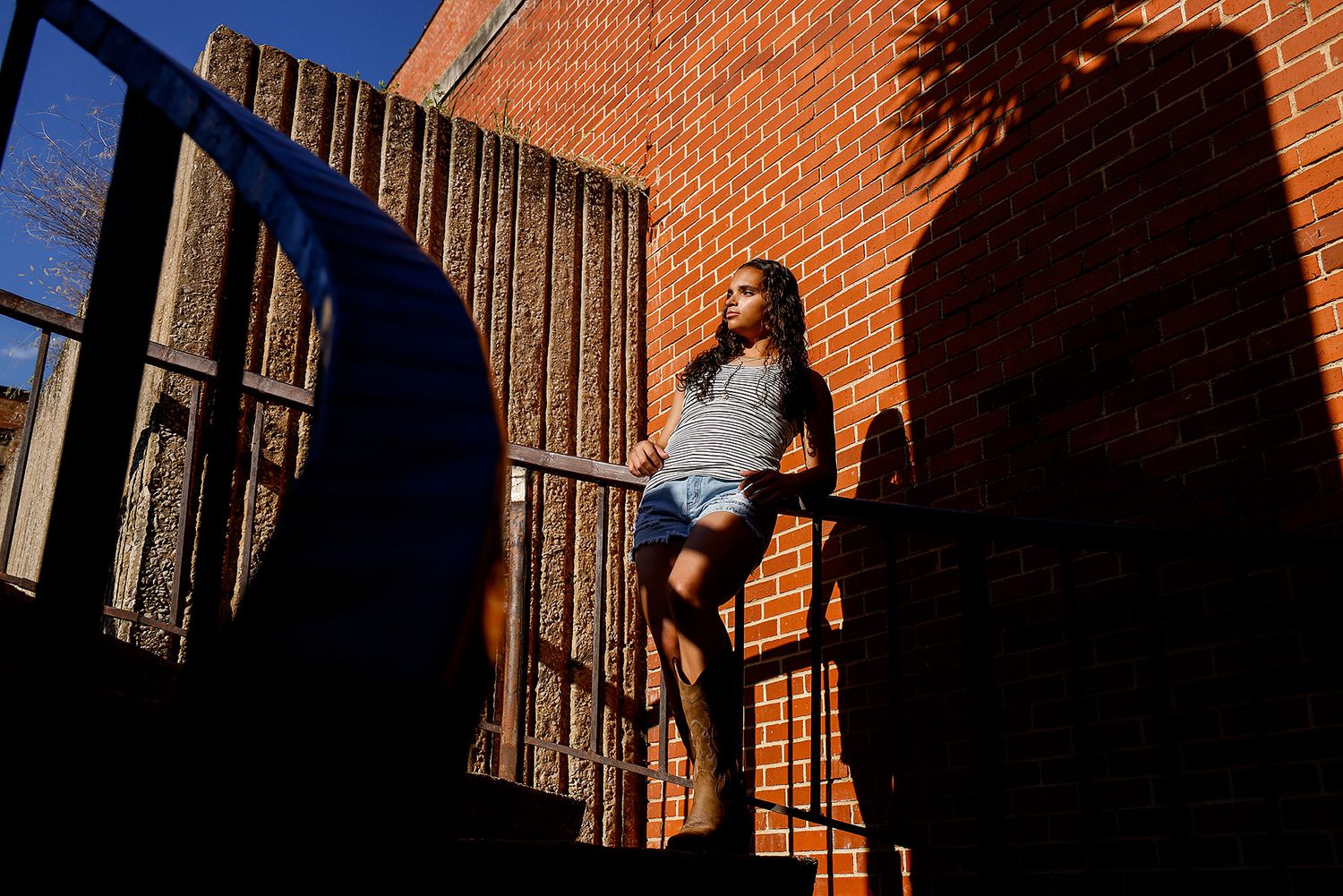 Young woman in denim shorts leans on stair railing against brick wall in dramatic sunlight and shadows.